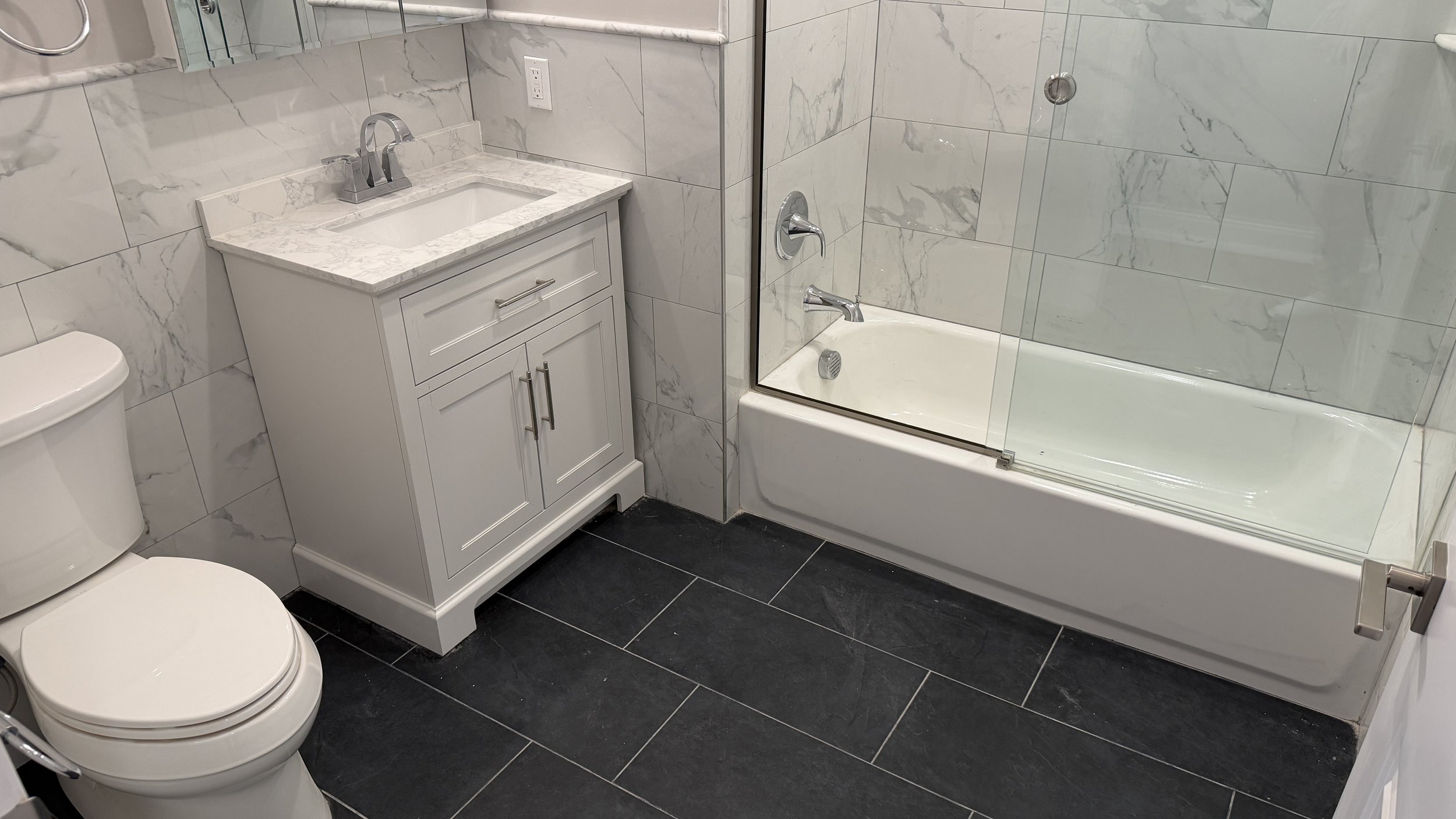 Bathroom with white vanity, marble countertop, toilet, bathtub with glass shower door, and marble tile walls.