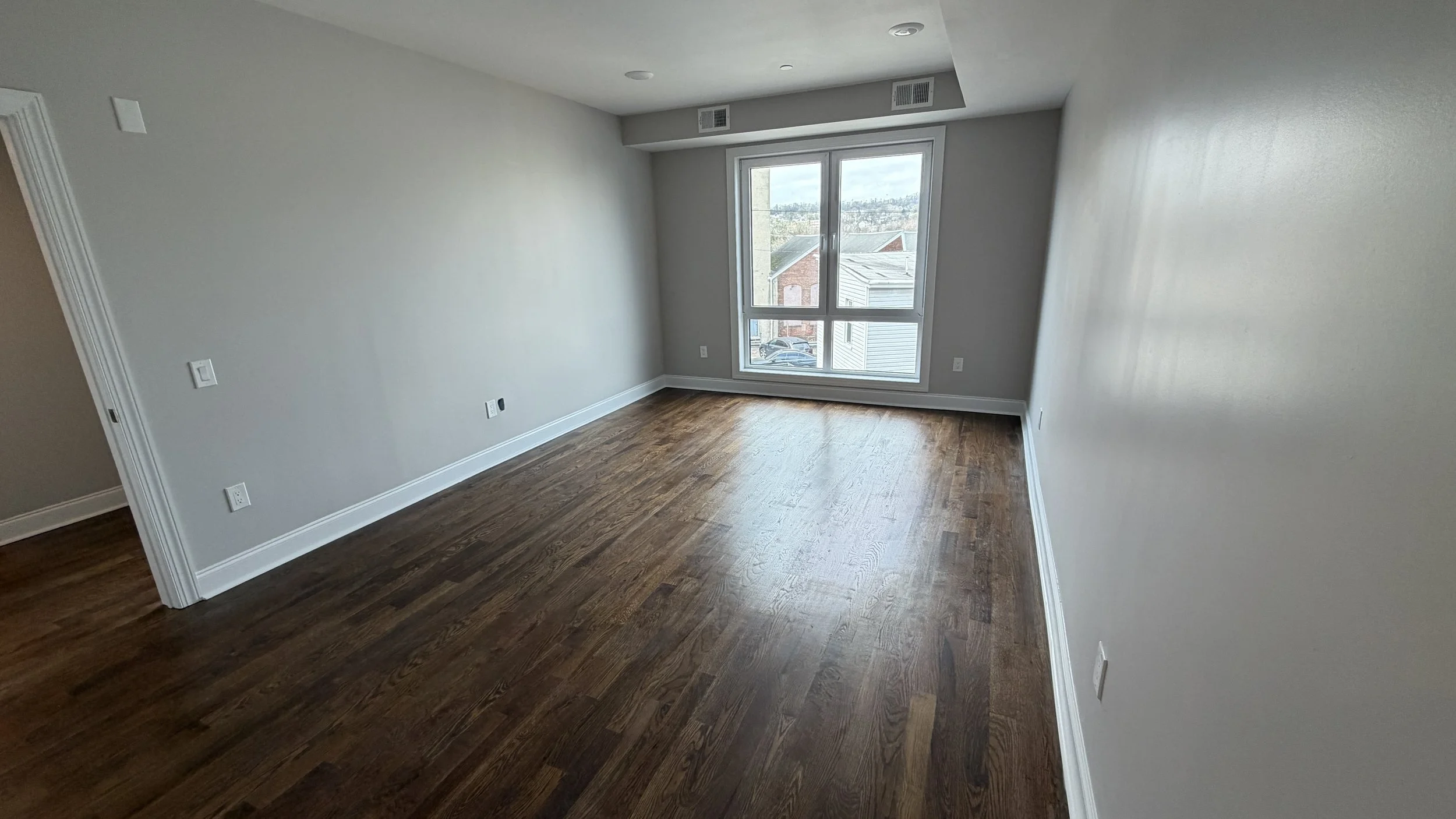 Empty room with hardwood floors, gray walls, and large window showing outdoor houses and sky.