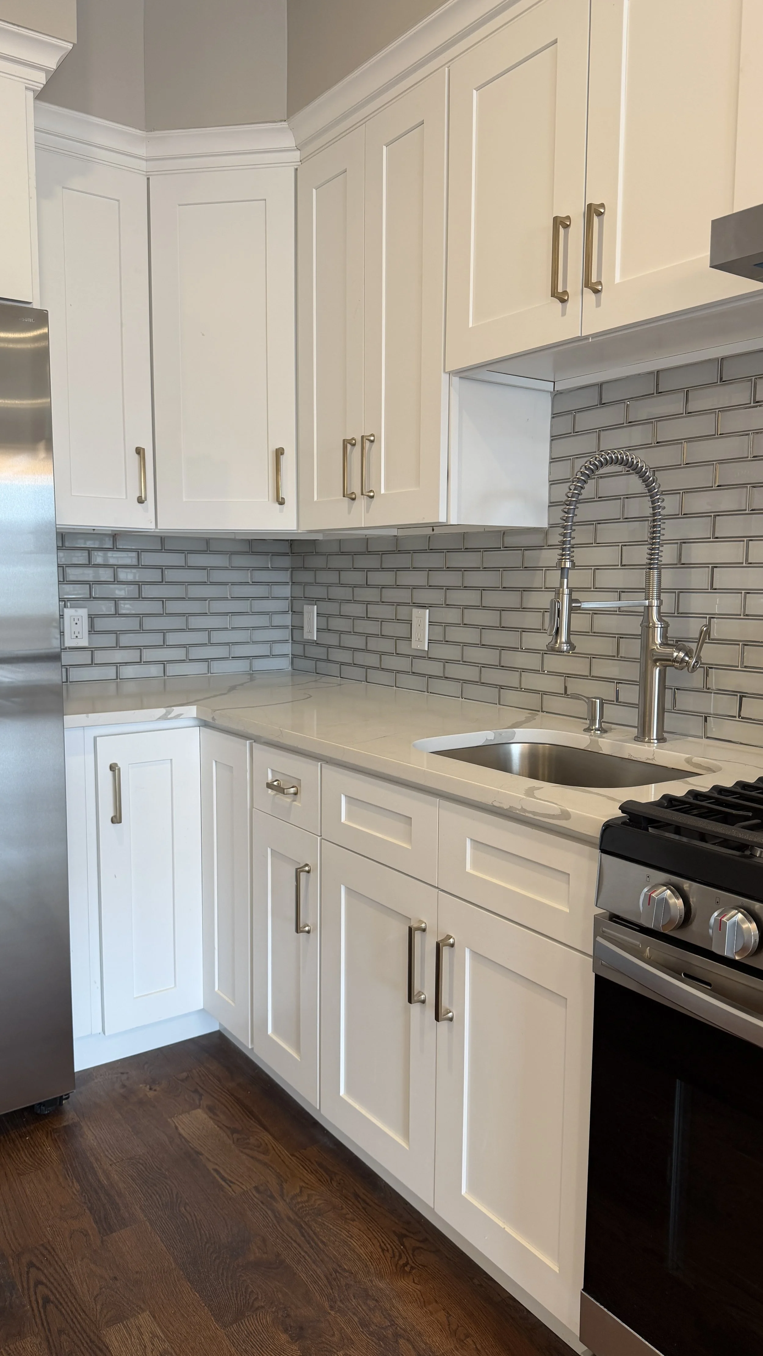 A kitchen with white cabinets, a gray subway tile backsplash, a stainless steel refrigerator, a white marble countertop, a small stainless steel sink with a high-arched spray faucet, and a black stove with oven.