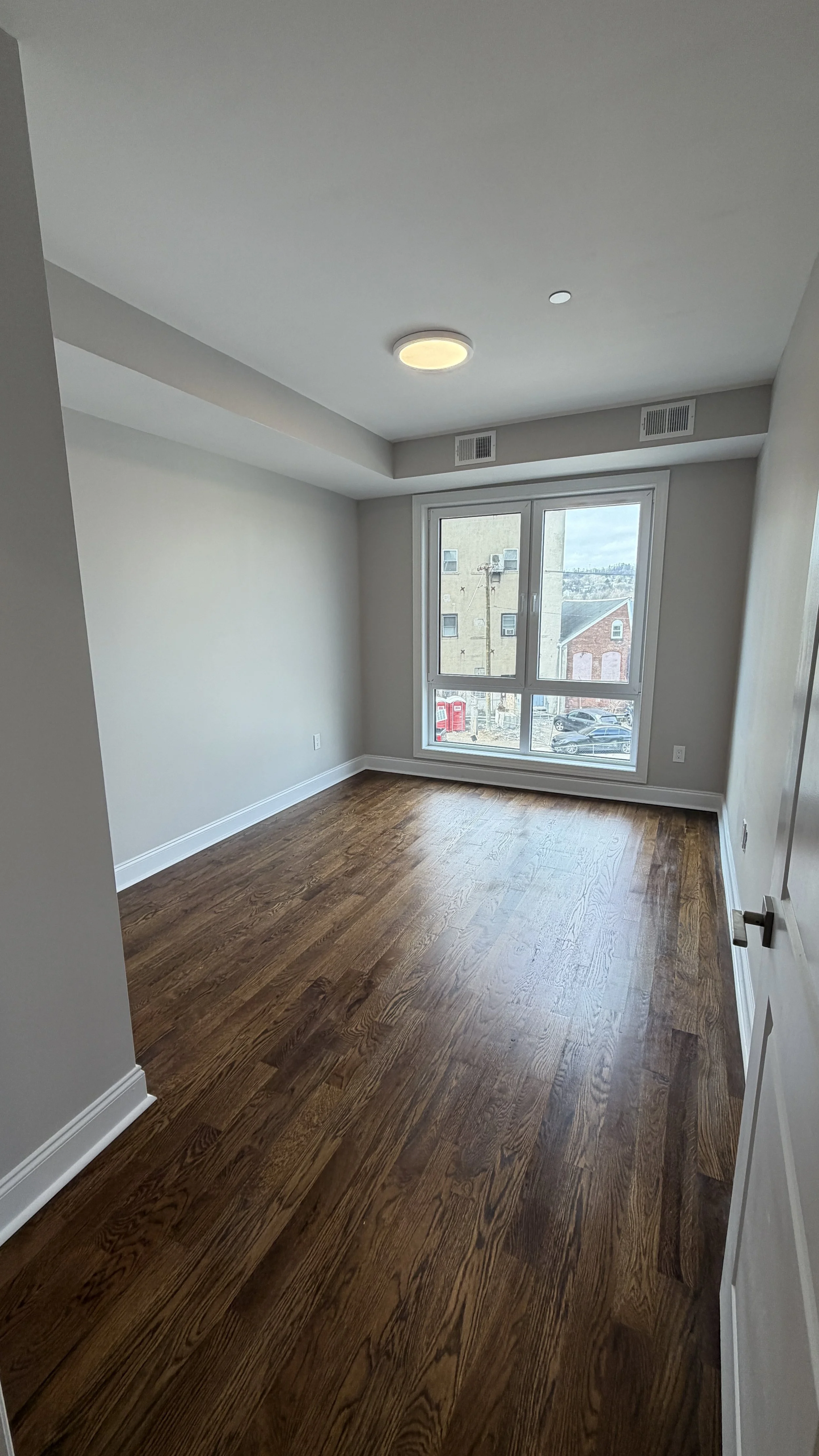 Empty room with wooden floors, beige walls, a large window, white baseboards, and a ceiling light fixture.