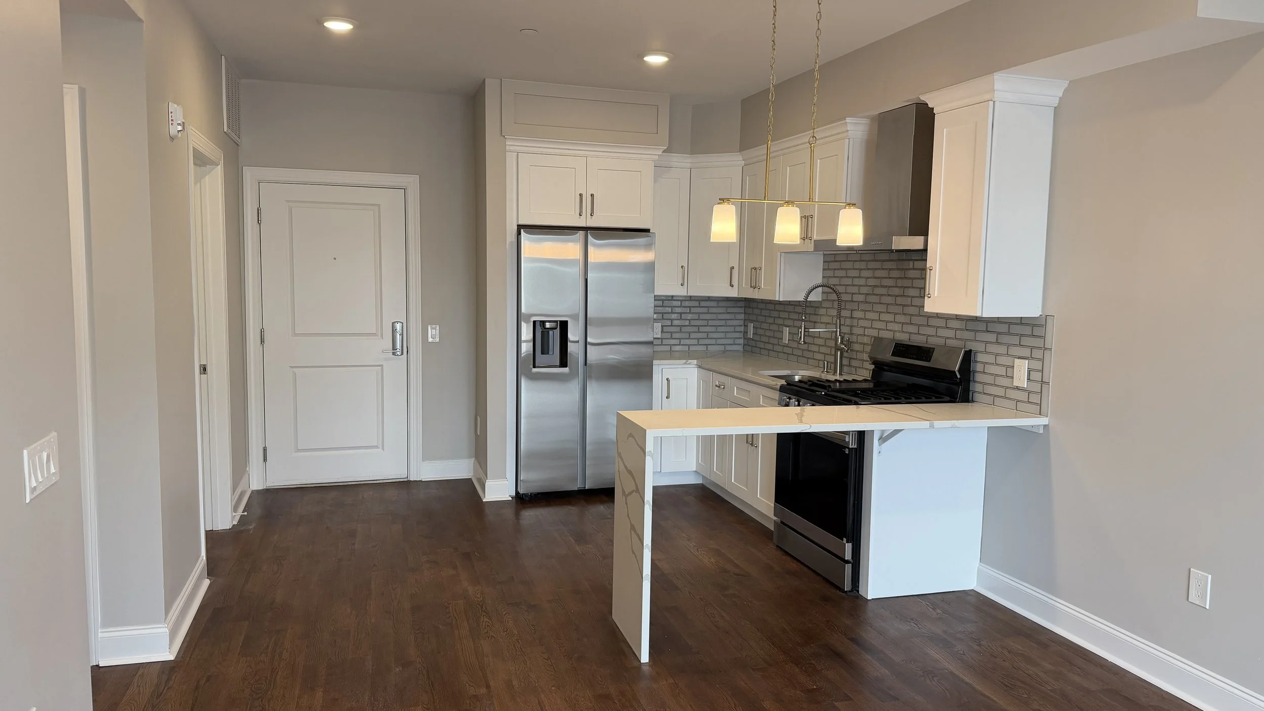 Modern kitchen with white cabinets, stainless steel refrigerator, black oven, gray subway tile backsplash, and hardwood floors.