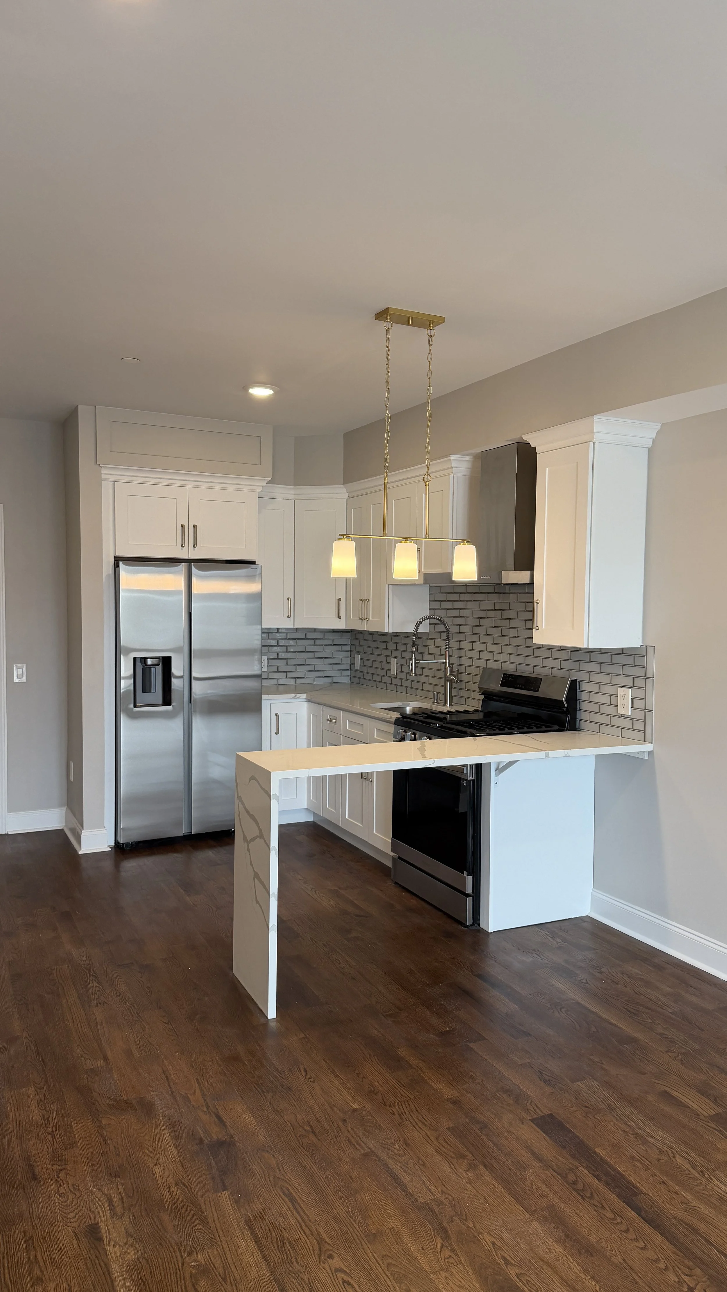 Modern kitchen with white cabinets, stainless steel refrigerator, black oven, gray backsplash, and wooden floor, featuring a hanging light fixture.