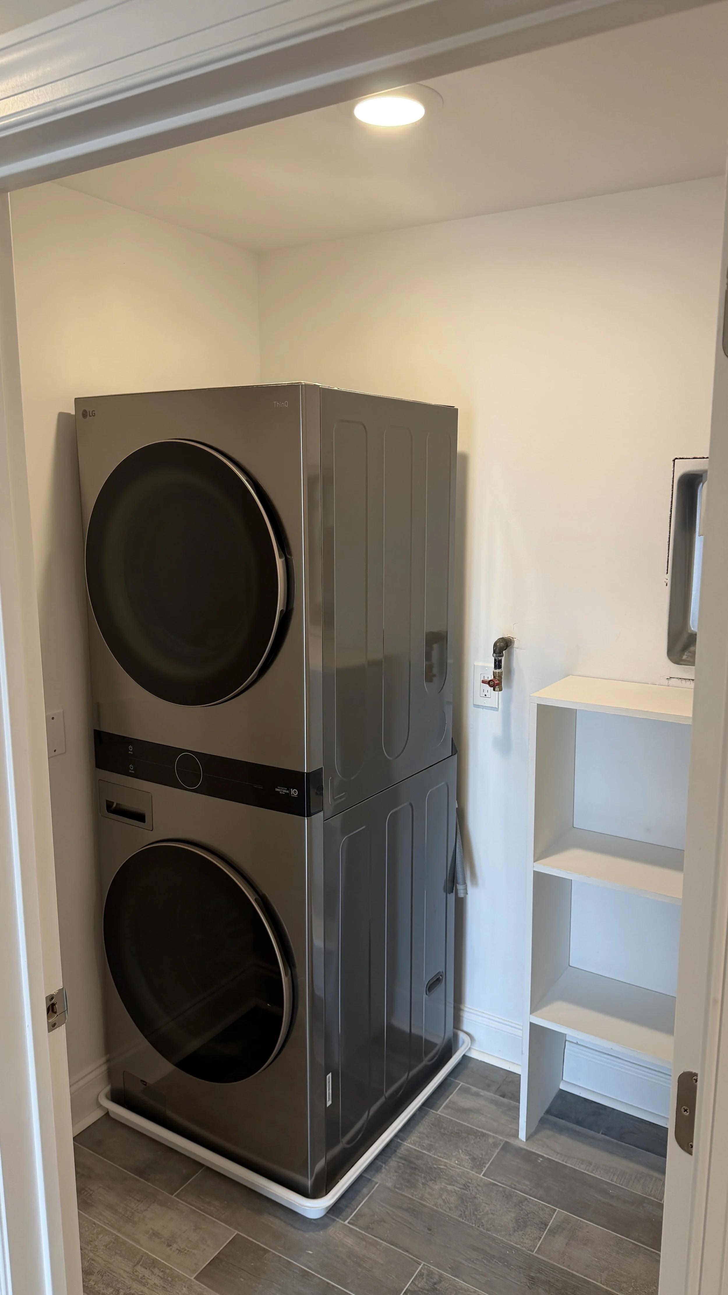 A laundry room with a stacked silver LG washer and dryer, a white shelf unit, and a gray tiled floor.