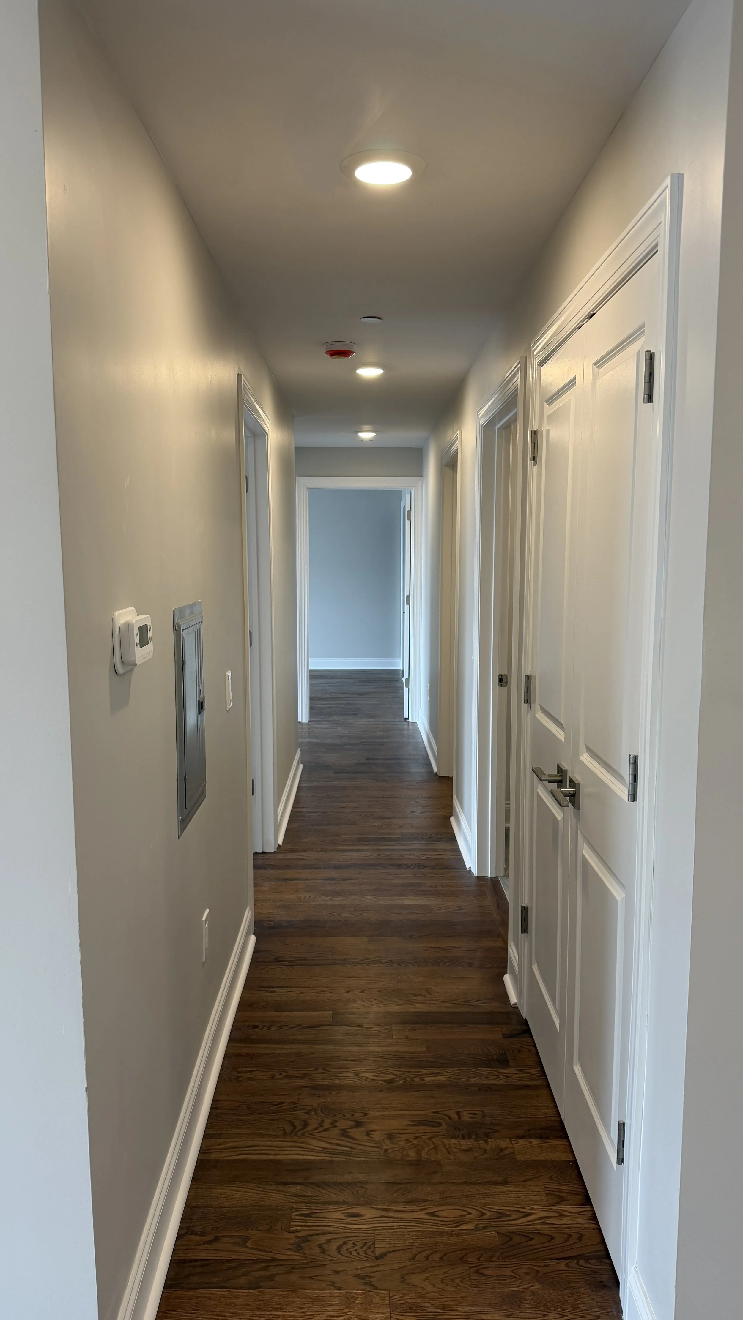 A hallway with white walls, wooden floor, and ceiling lights; doors along the corridor and a thermostat on the wall.