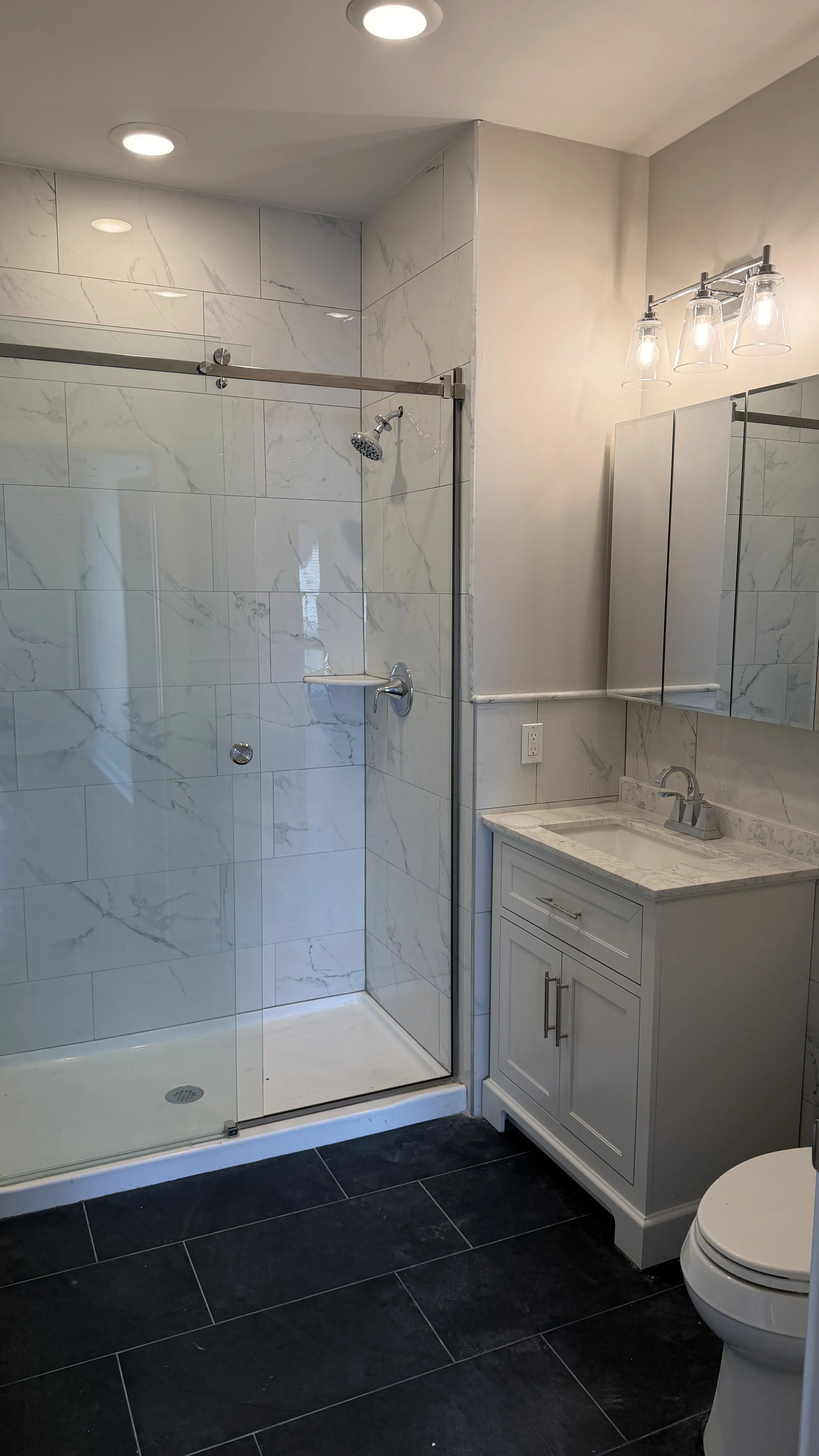 Modern bathroom with a glass-enclosed shower, white marble tiles, a small white vanity with a marble countertop and a mirror, and a toilet, with dark floor tiles and ceiling lights.