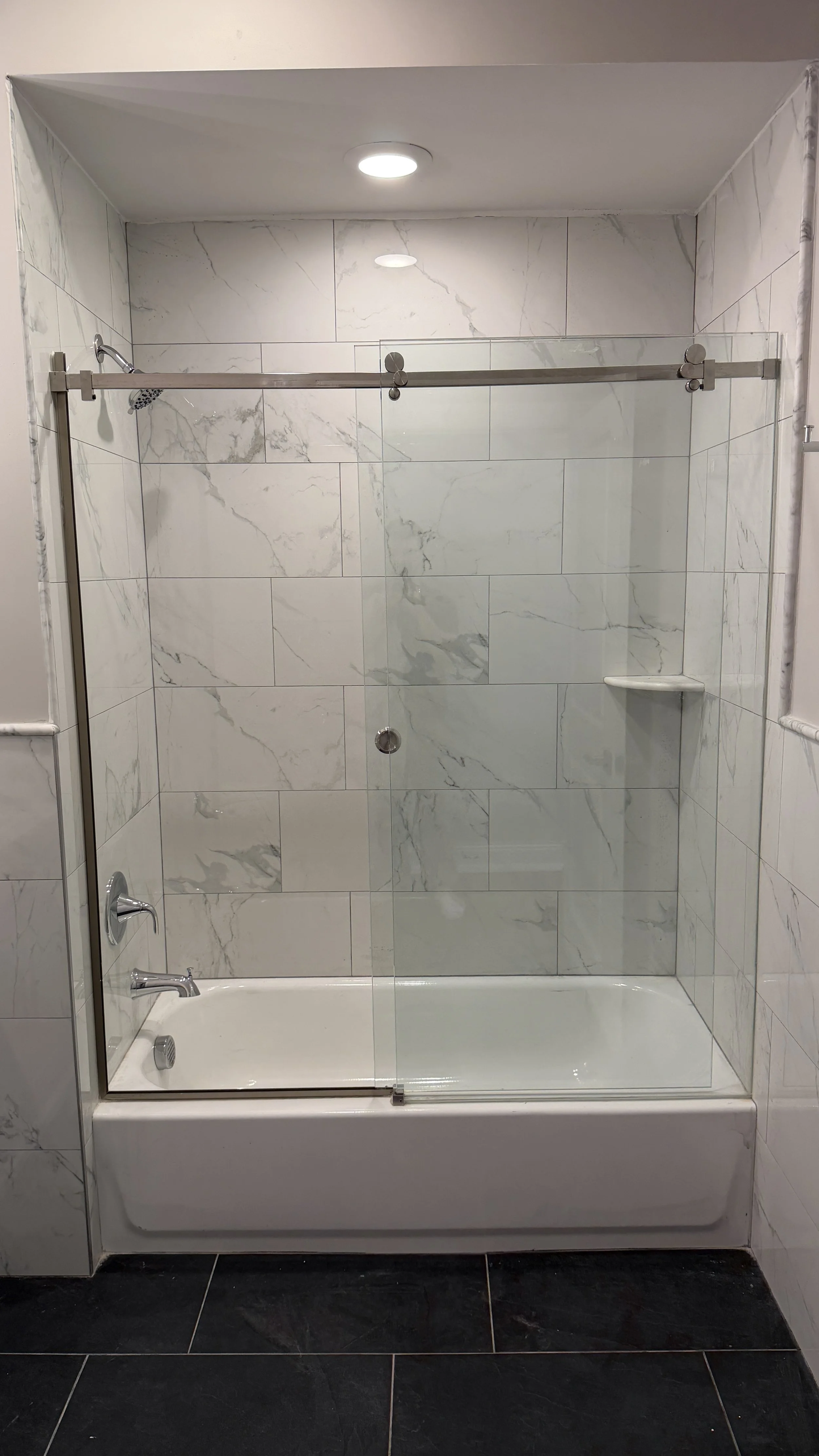 A modern shower with white marble tiles, a glass sliding door, a metal towel bar at the top, and a built-in corner shelf. The shower has a showerhead, a faucet, and a bathtub below. The walls are tiled and the floor is black with tiles.