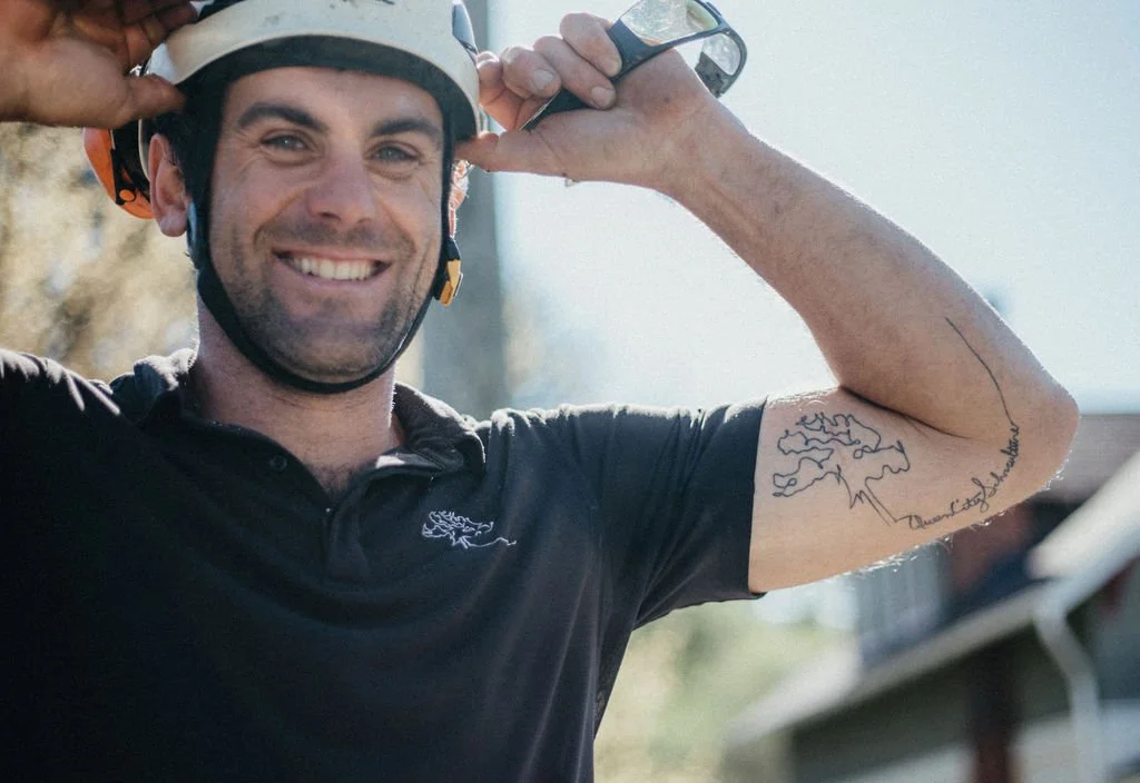 Smiling man in a black polo shirt and helmet, adjusting his helmet strap, outdoors on a sunny day.