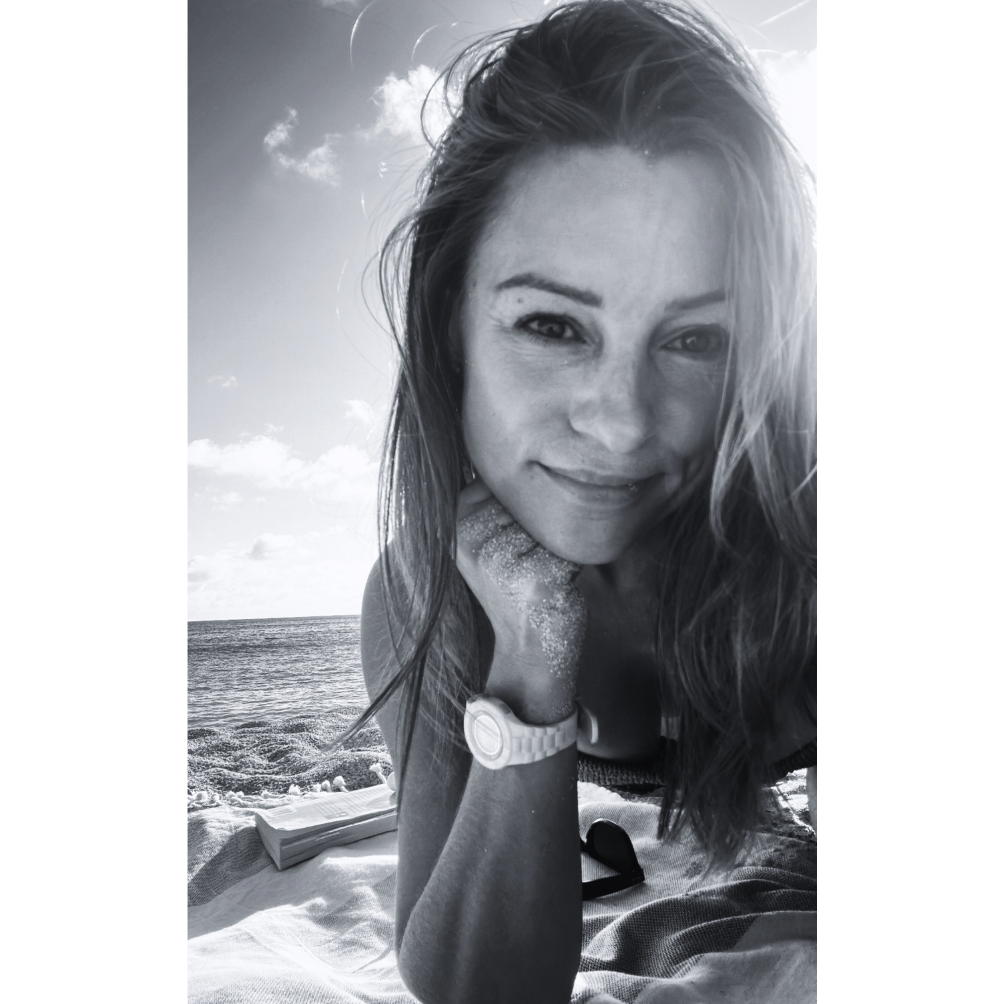 A black and white photo of Sloan Lauinger at the beach in St Barth on Gouverneur Beach, just out of the water with her RayBan sunglasses and a book beside her.