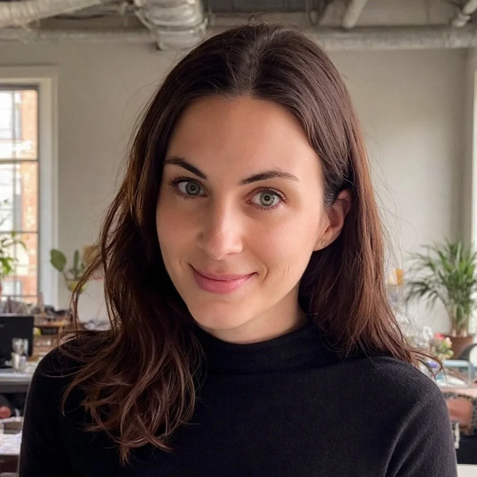 A woman with dark brown hair and blue eyes smiling indoors, wearing a black top in a room with large windows and plants.