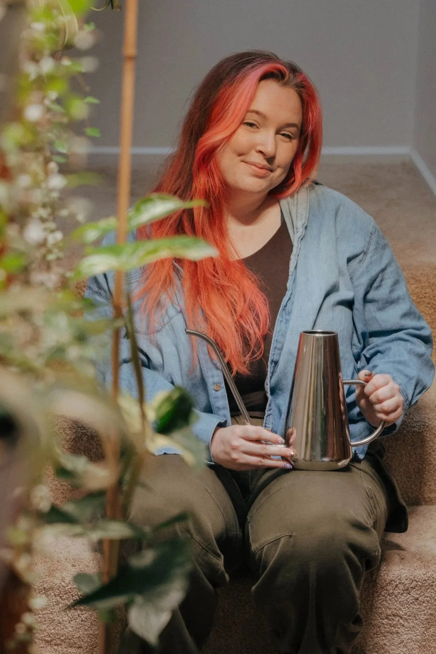 A woman with red hair, wearing a black shirt and a light blue denim jacket, sitting on a carpeted floor, holding a metallic watering can, smiling softly at the camera. A leafy green plant is partially visible in the foreground.