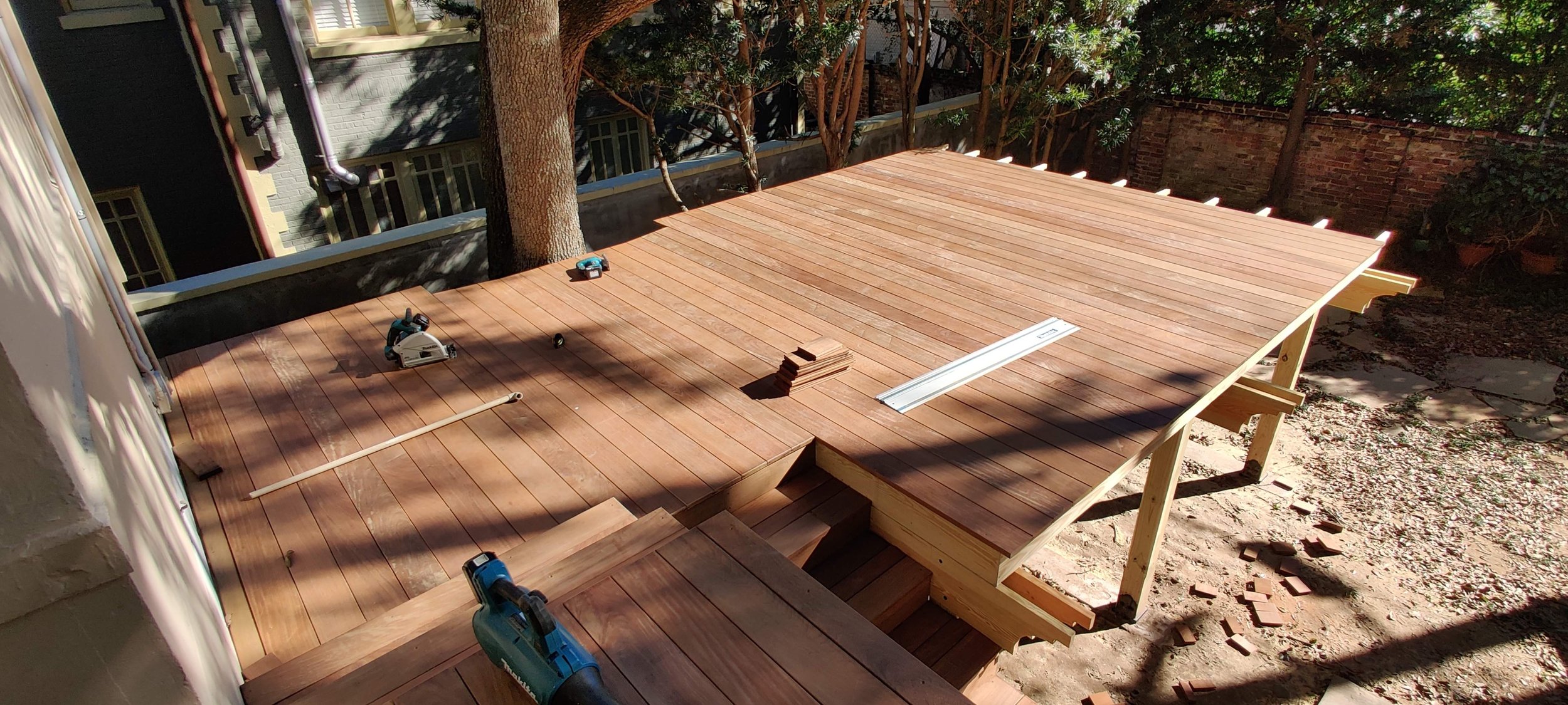 The image shows a wooden deck under construction in a backyard with trees and a brick wall. Construction tools and materials, including a circular saw, measuring tape, wooden planks, and small stacks of wood, are scattered on the deck. The decking is