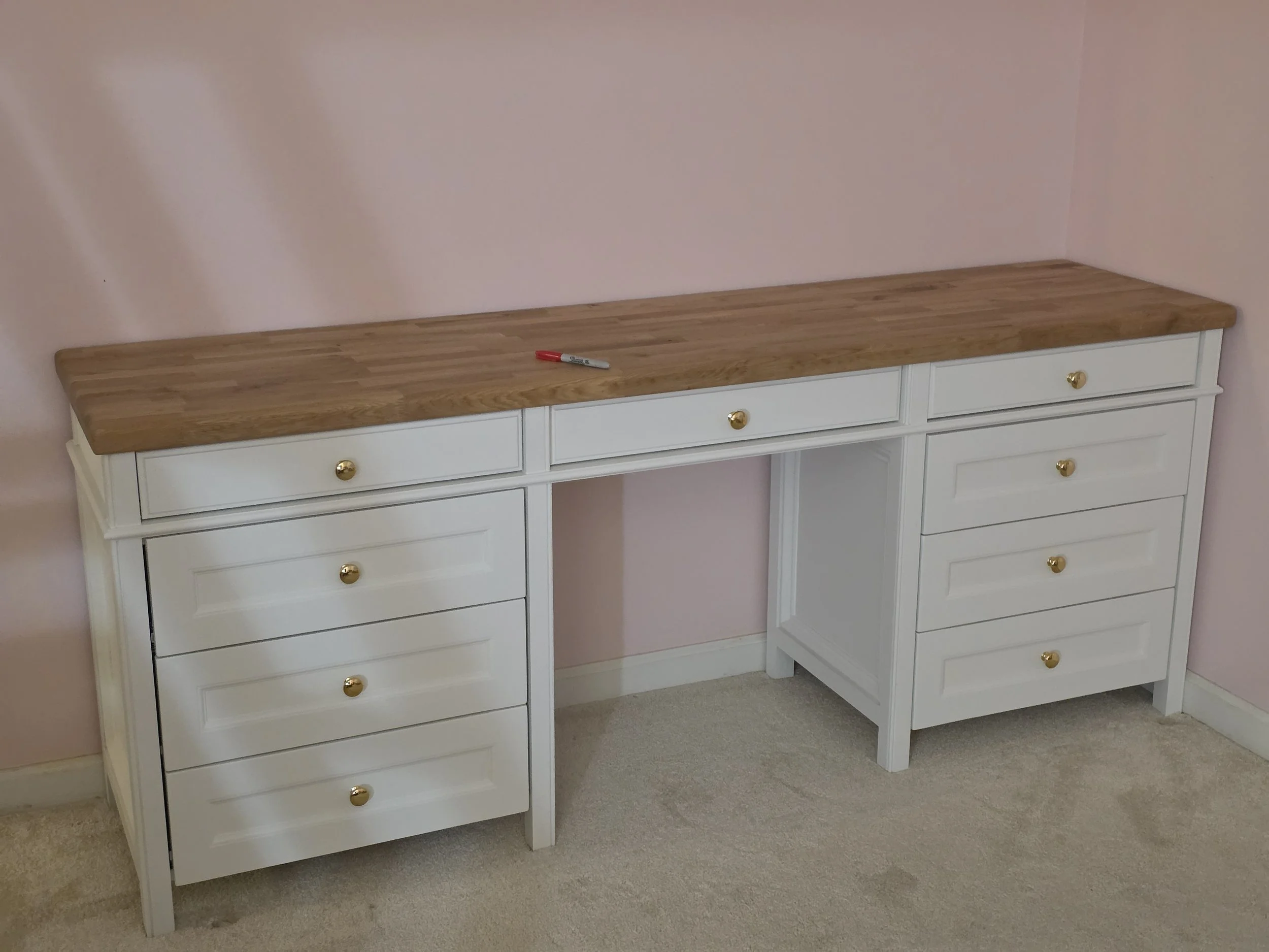 White wooden desk with five drawers and a light brown wooden top, positioned against a pink wall with beige carpet.
