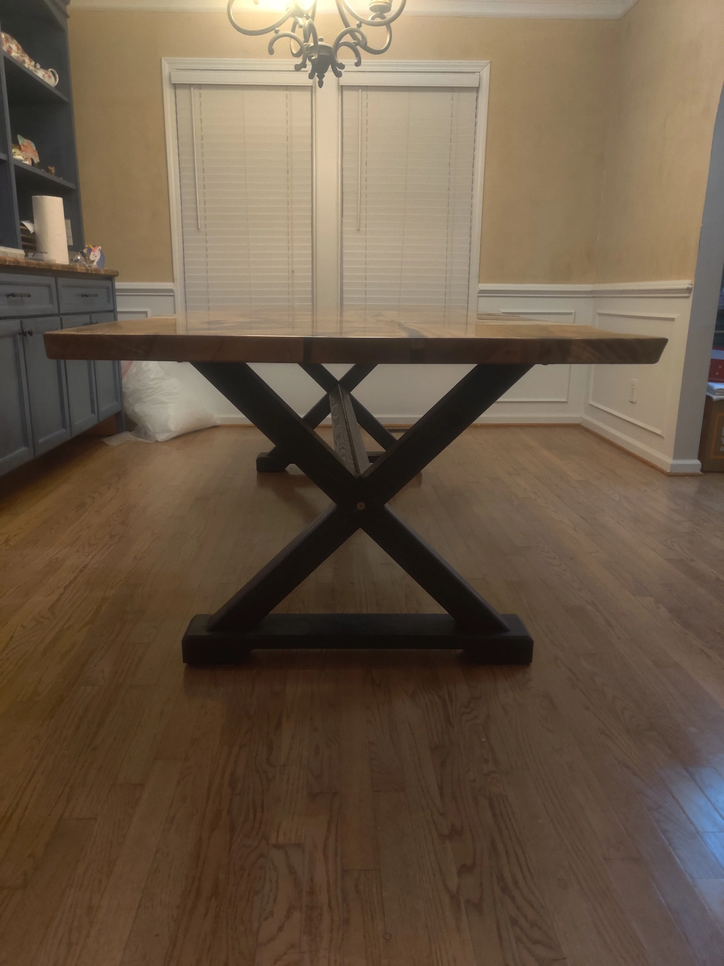 Wooden dining table with a dark X-shaped pedestal base in a dining room with hardwood floors, white paneled walls, and a chandelier.