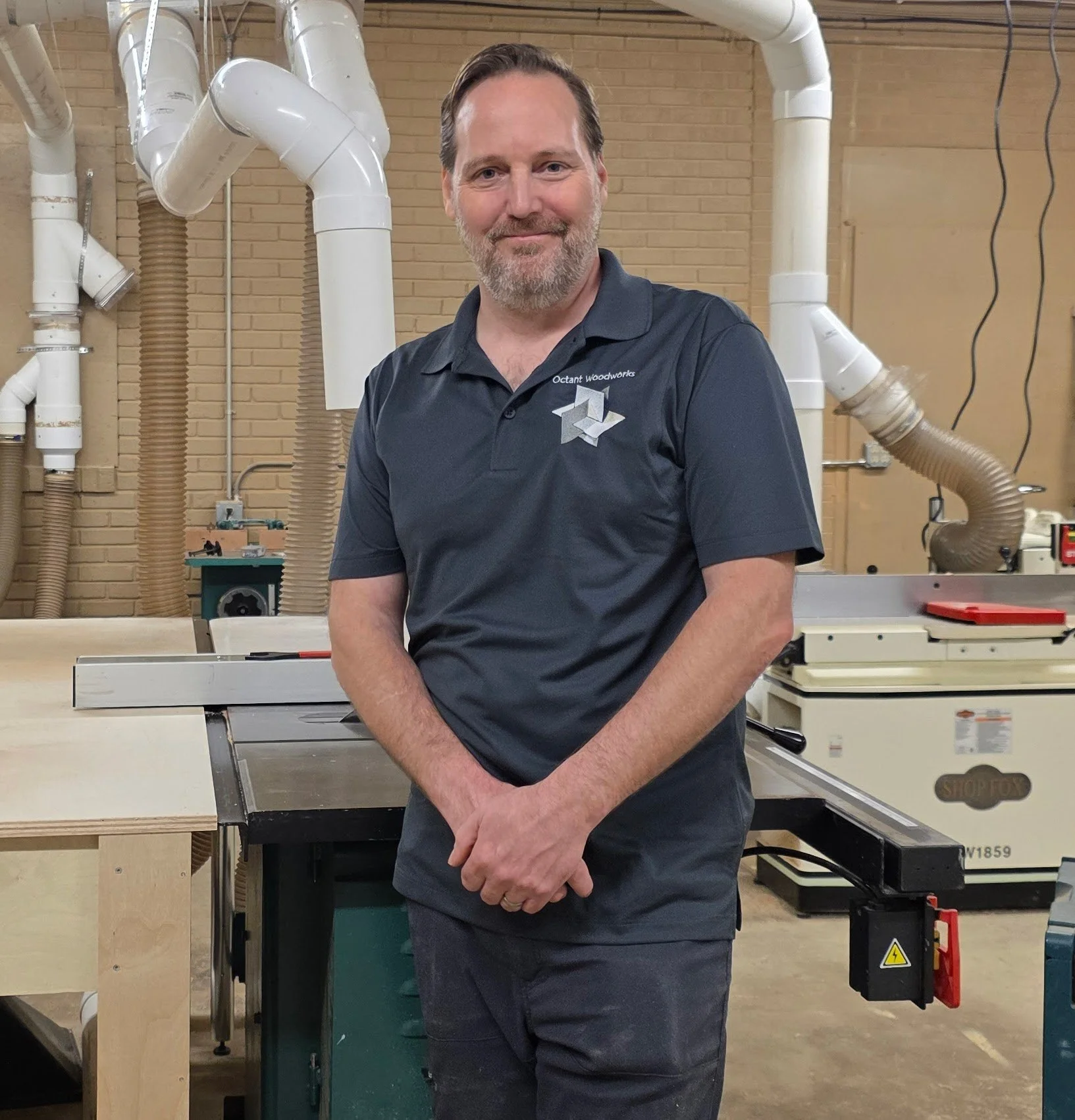 Chris Harmon, standing in a woodworking shop with woodworking tools and machinery in the background, wearing a dark polo shirt with a logo that reads 'Octant Woodworks'.
