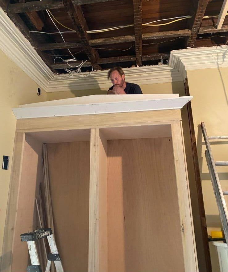Chris Harmon inspecting the ceiling of a room while standing on a built-in wooden cabinet under construction, with exposed ceiling beams and wiring.