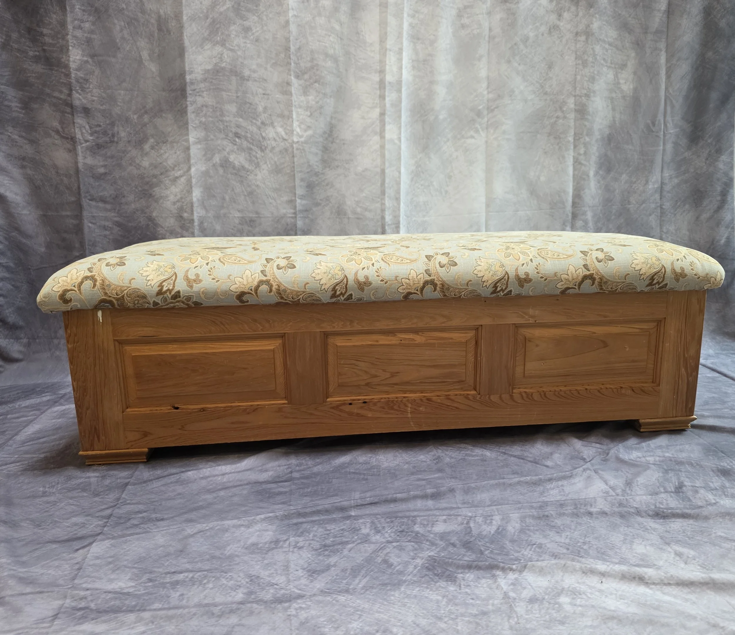 A wooden bench with a cushioned floral-patterned top against a gray backdrop.