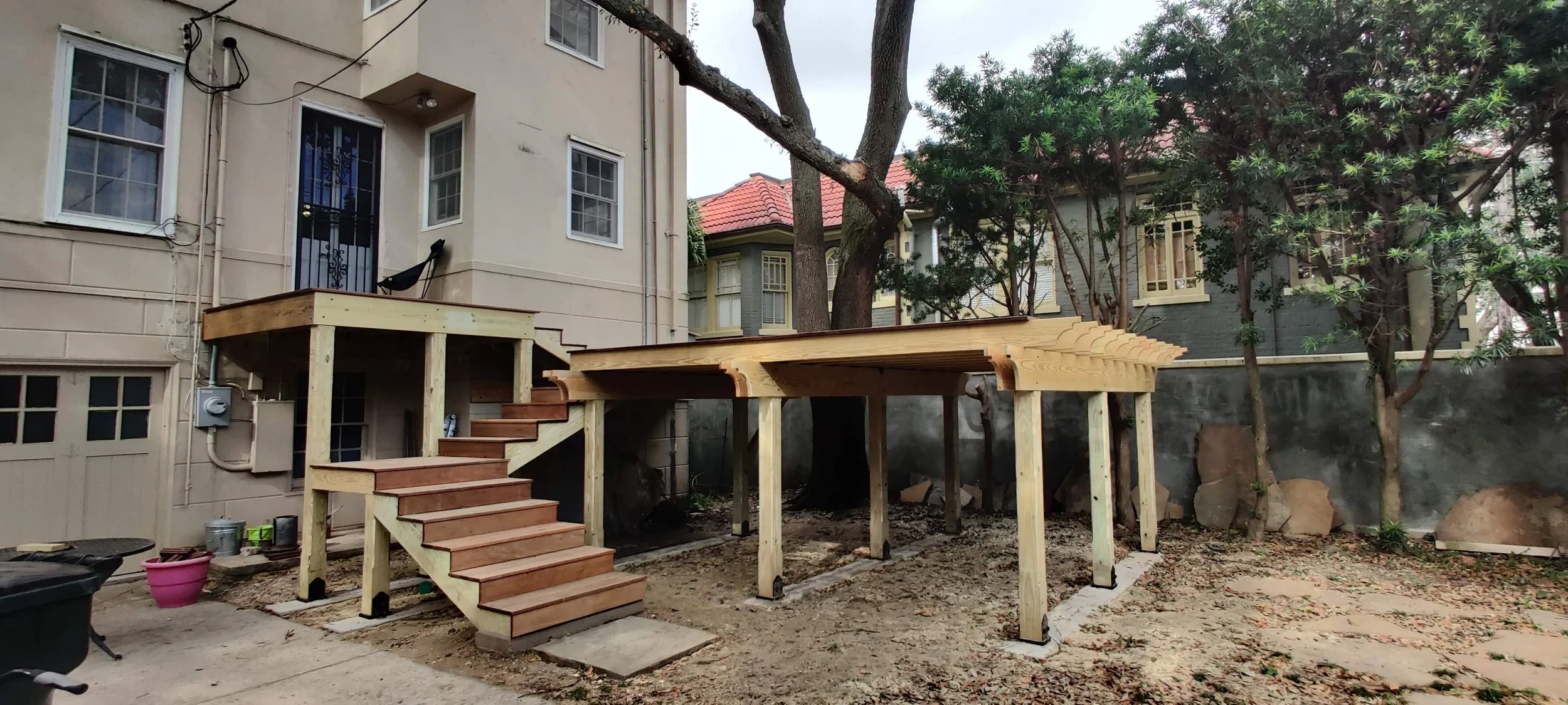 Construction of a wooden deck with stairs in a backyard, surrounded by trees and neighboring houses, with dirt ground and construction materials nearby.