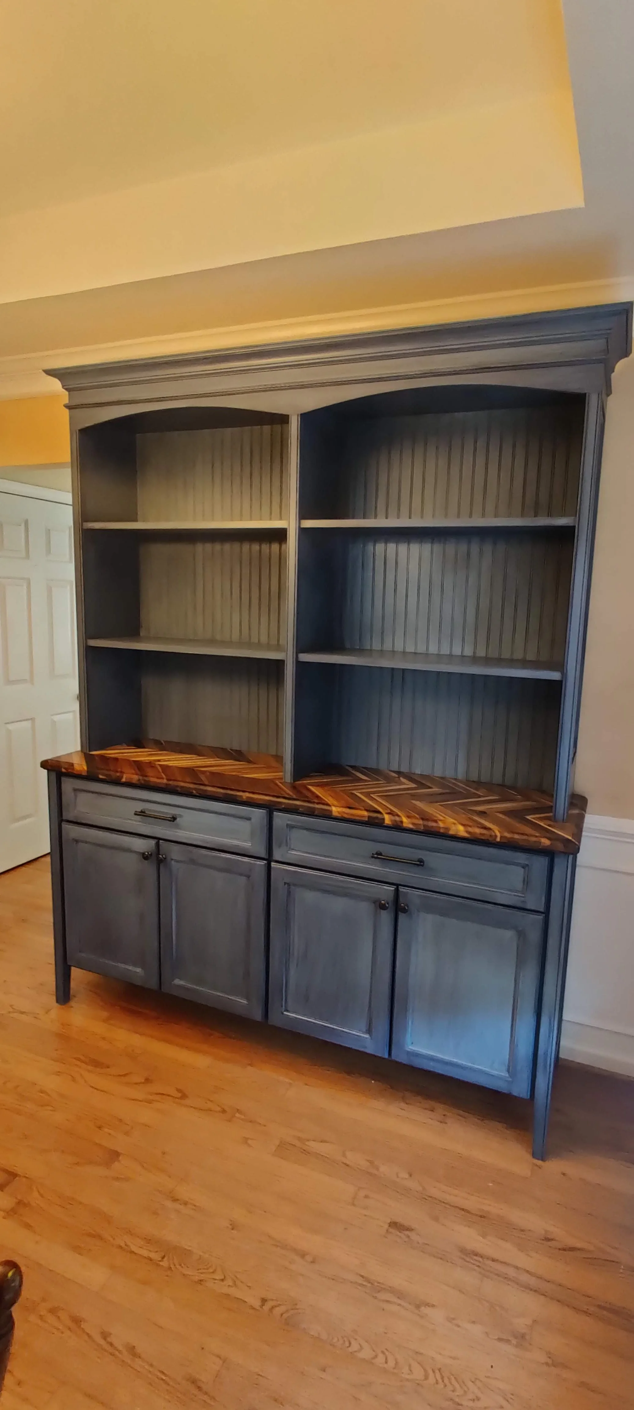 A wooden hutch painted in distressed gray with a polished wood countertop, featuring open shelves and closed cabinets beneath, situated on a hardwood floor in a room with cream-colored walls.