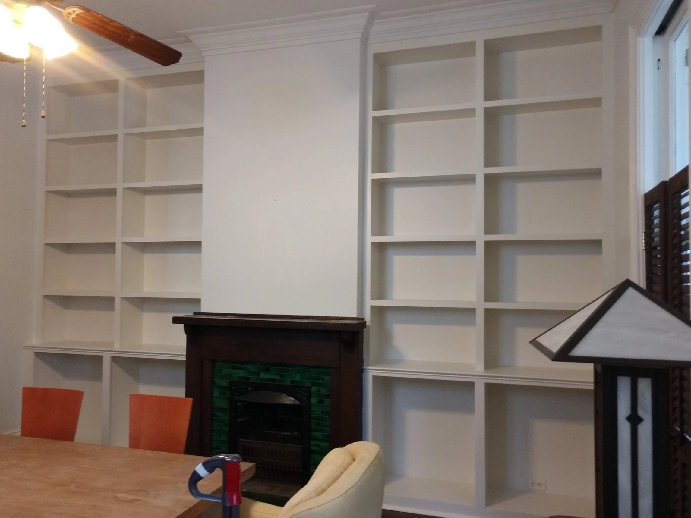 Living room with white built-in bookshelves and cabinets, a fireplace with green tiles, a wooden table, and chairs, with a ceiling fan and window blinds.