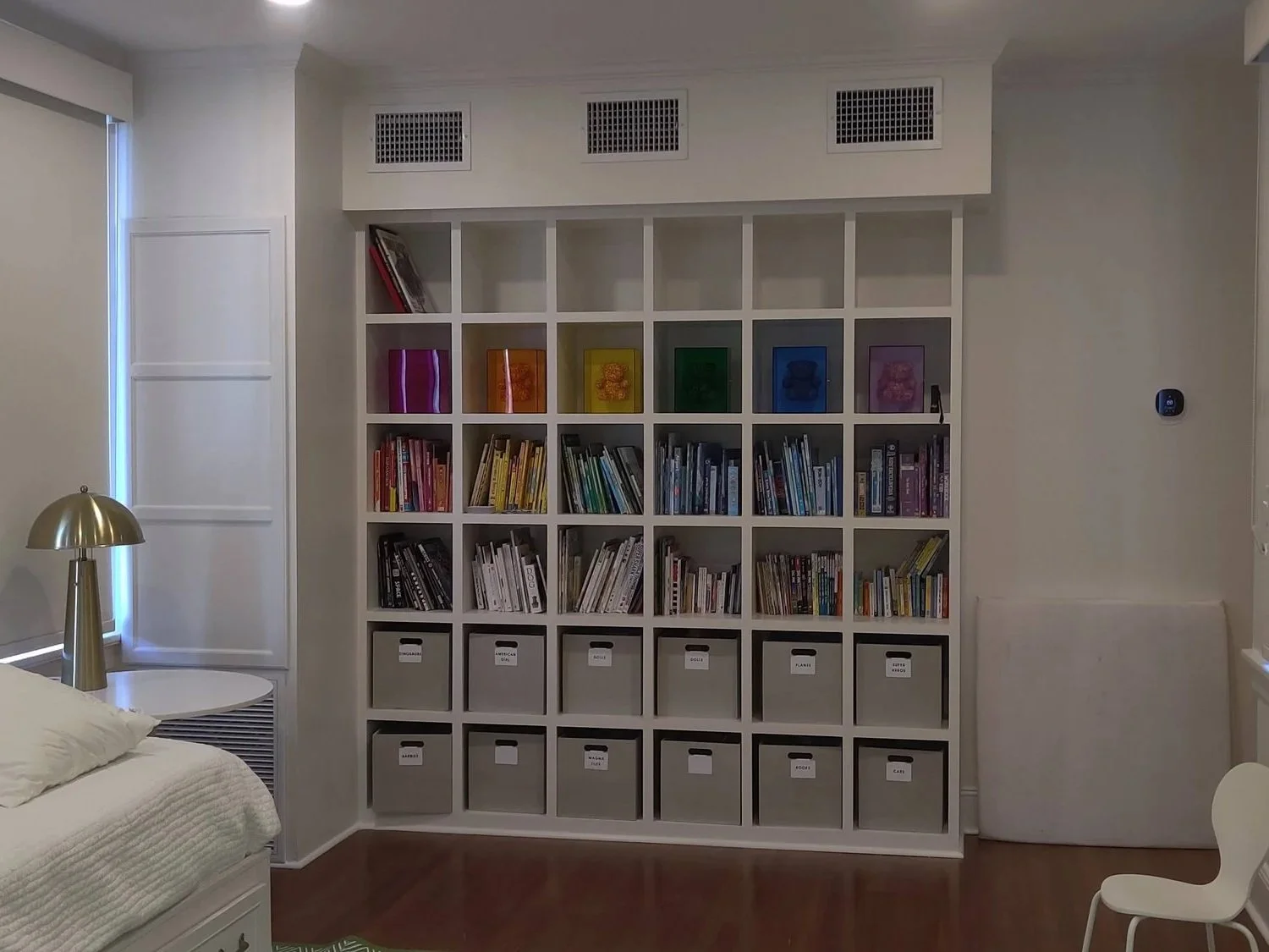 Custom built white shelving unit filled with books and colored storage bins in a room with a side table, a lamp, and a white bed.