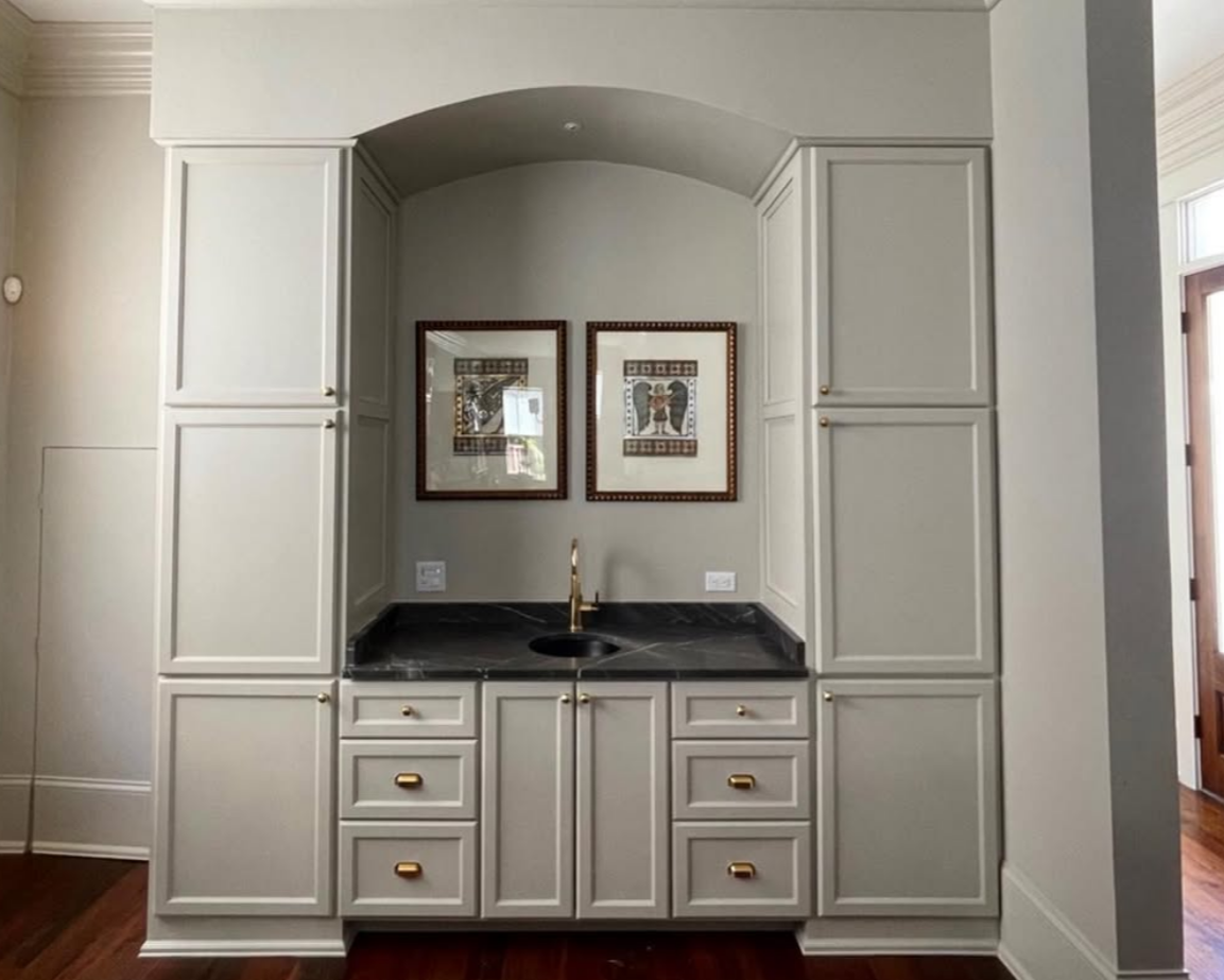 Custom built-in wet bar with white cabinetry, black marble countertop, a small sink, gold handles, and two framed art pieces on the wall