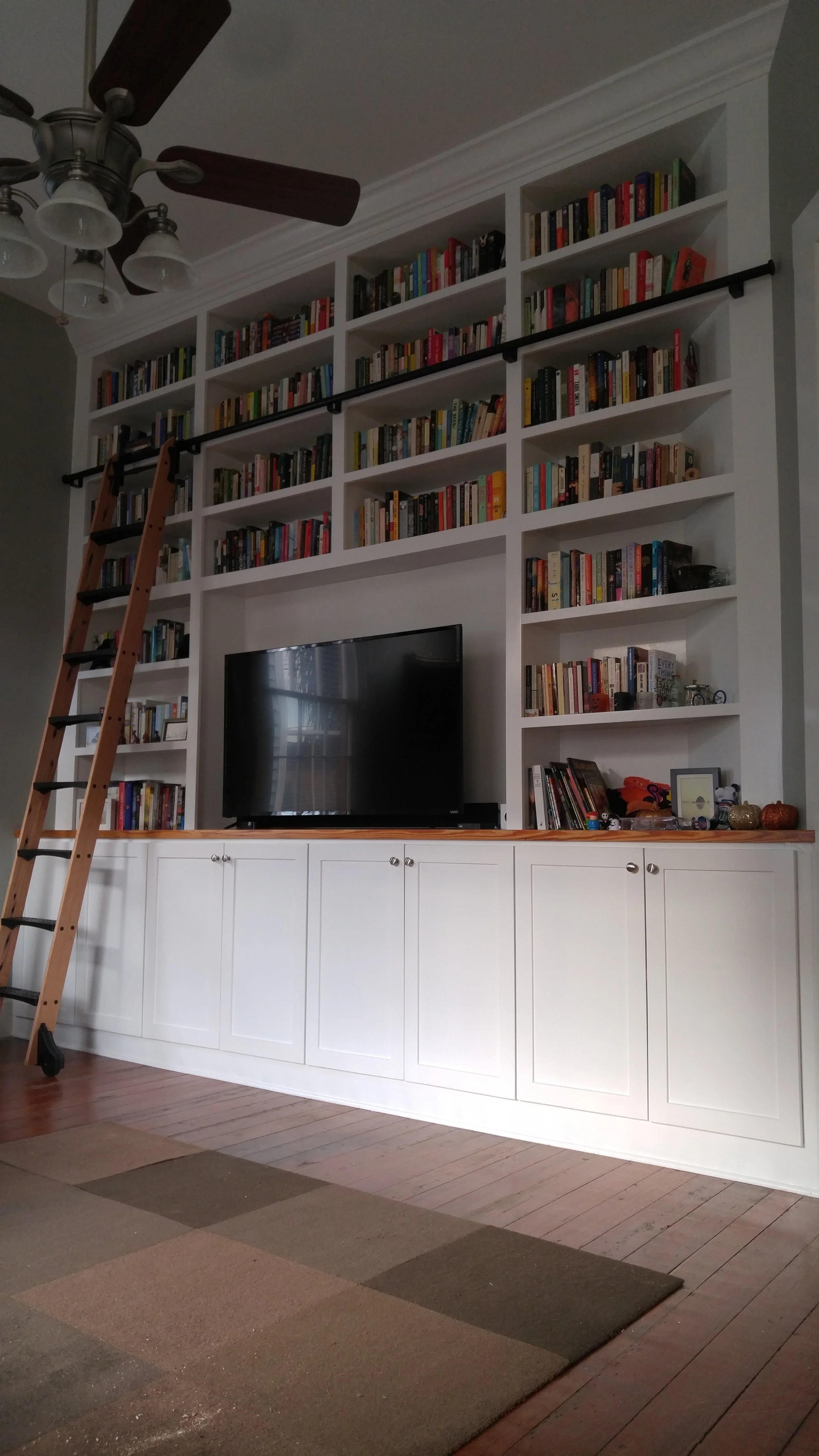 A custom built-in white entertainment center with multiple shelves filled with books, a black TV, a rolling library ladder leaning against the bookshelf, a rug on a wooden floor, and a ceiling fan with lights.