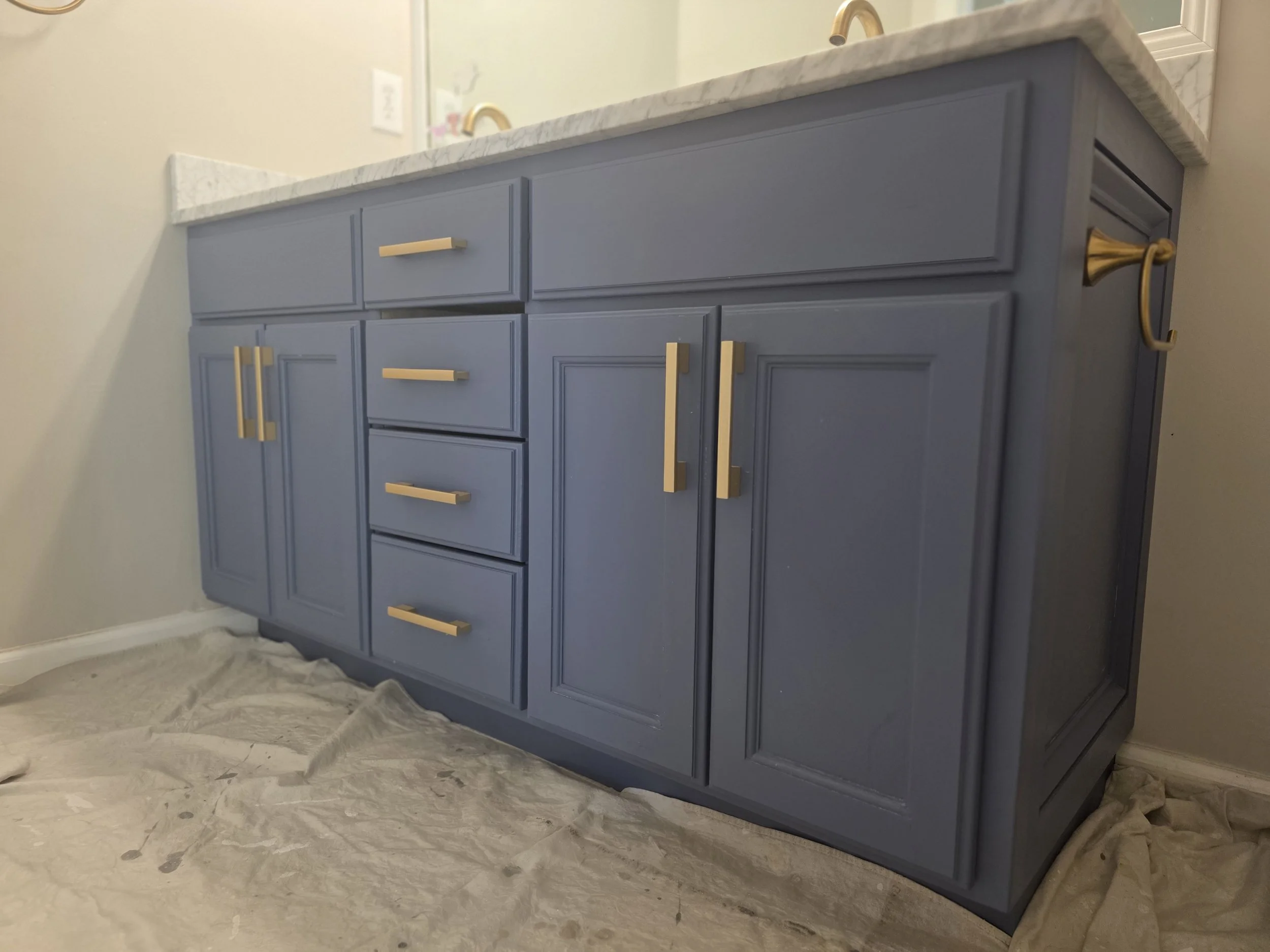 custom built blue bathroom vanity with gold handles and a marble countertop, positioned against a light colored wall with a mirror above.