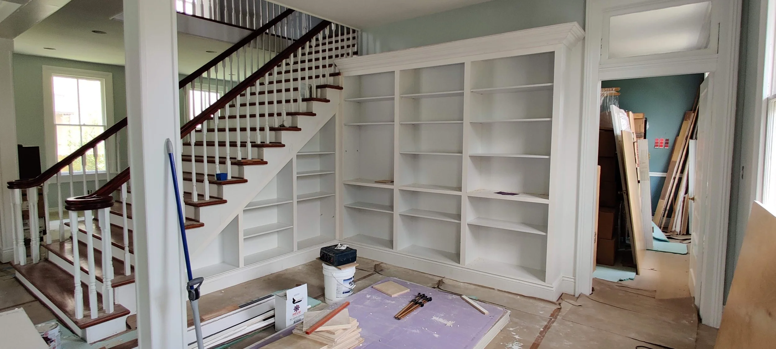 Interior of a house under renovation showing a white built-in bookshelf, a staircase with dark wood treads and white risers, and construction materials scattered on the floor.