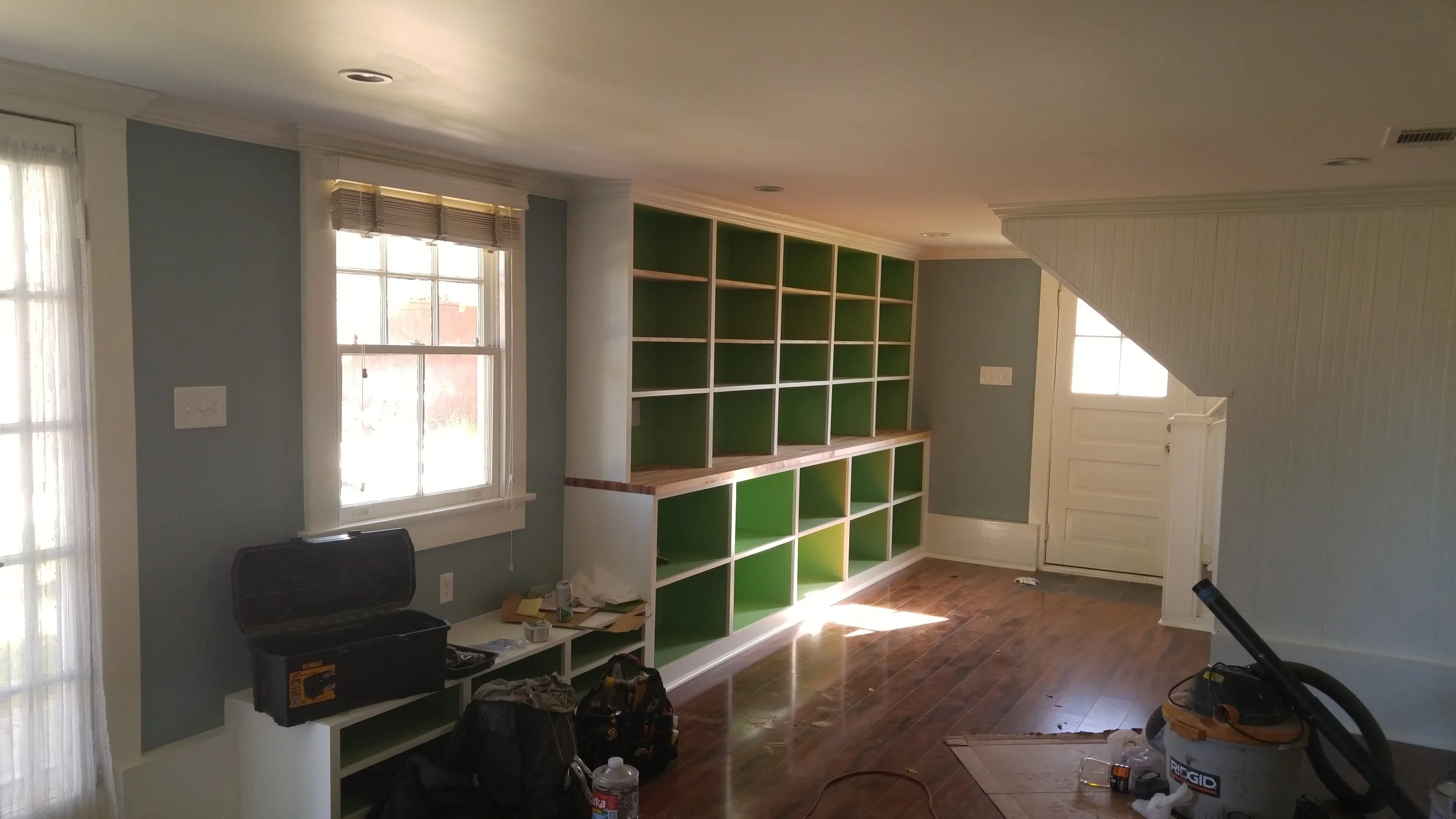 Room with light blue walls, large window with blinds, built-in shelf unit with green interior, and hardwood floor. Tools and supplies are on the floor and furniture, indicating ongoing installation or renovation.
