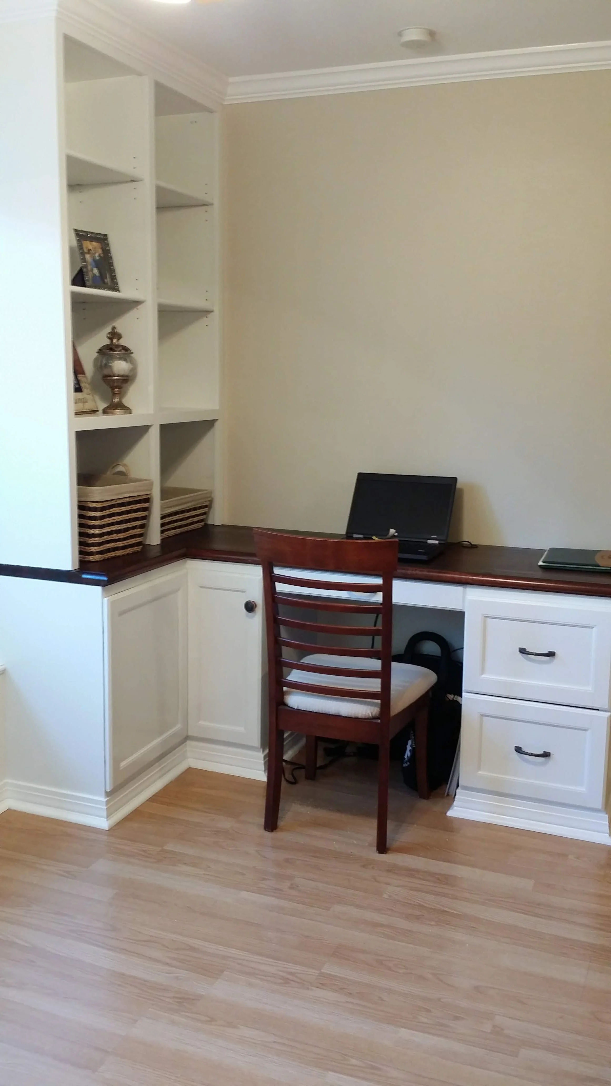 Home office corner with built-in white cabinetry, a wooden desk with a closed laptop, a wooden chair with a white cushion, and decorative items on the shelves, including a framed photo and a glass jar.