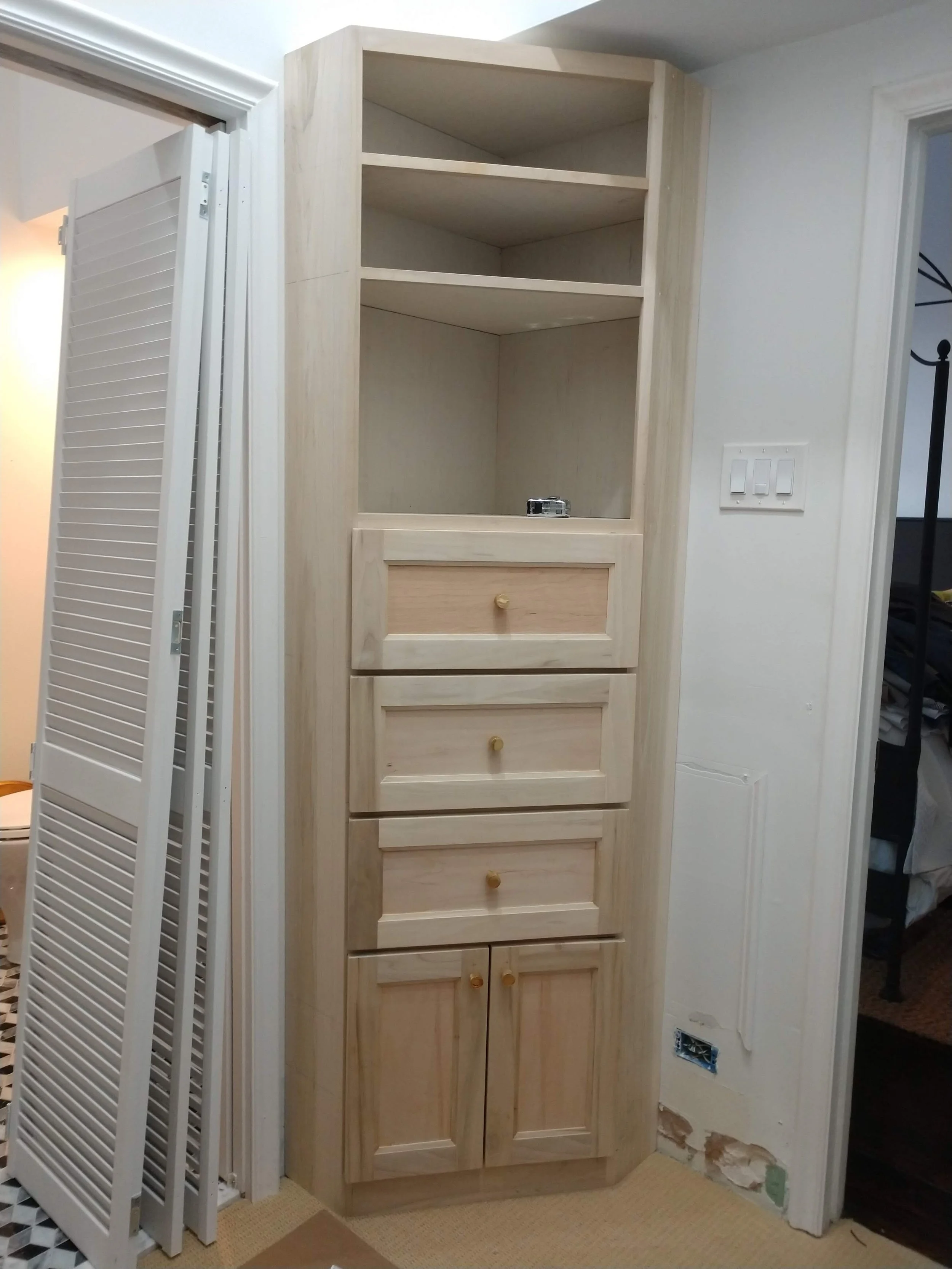 Unfinished tall wooden cabinet with open shelving and four drawers, situated in a room with white walls.