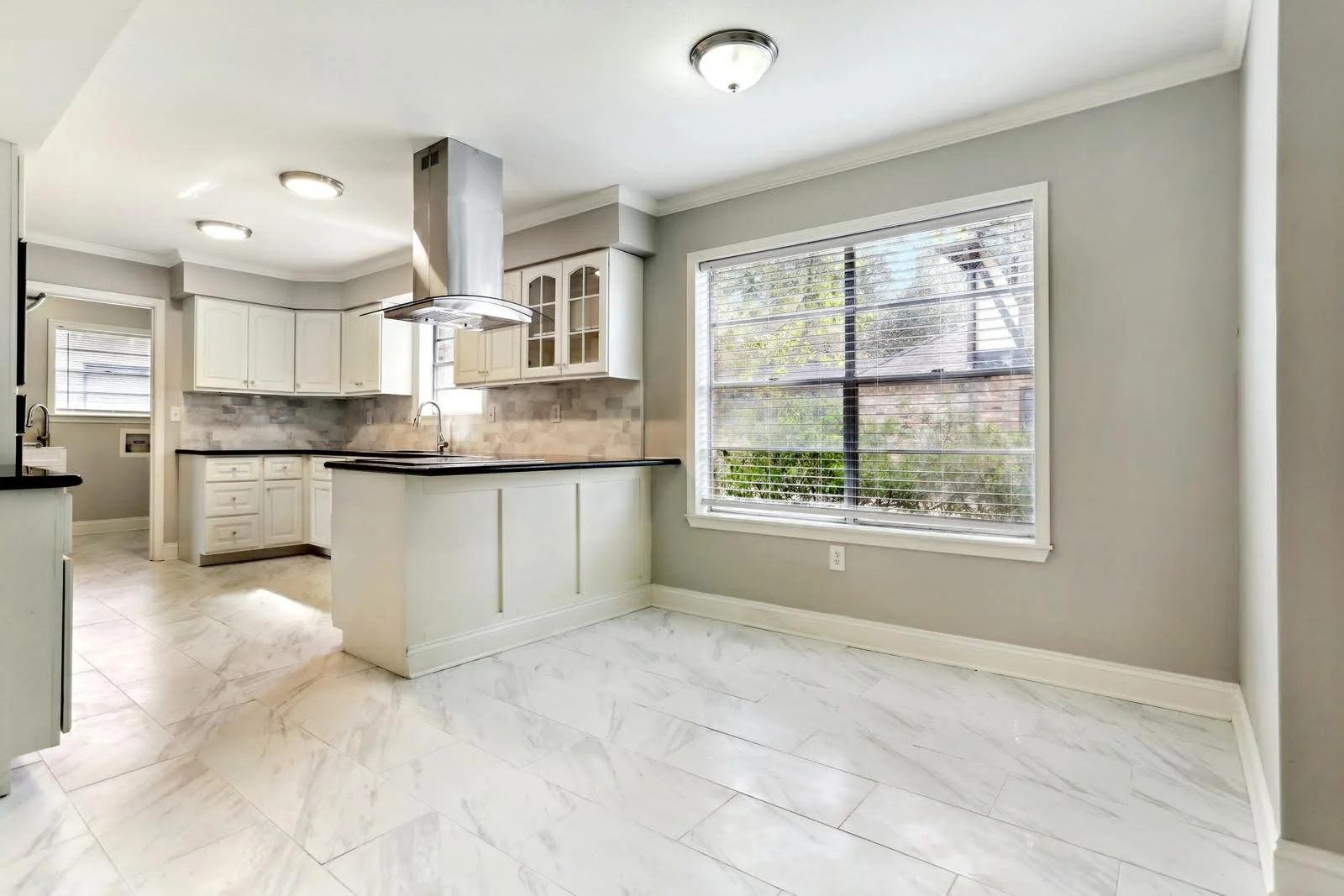 custom kitchen with white cabinets, black countertops, large window with blinds, marble floor, and a gray backsplash.