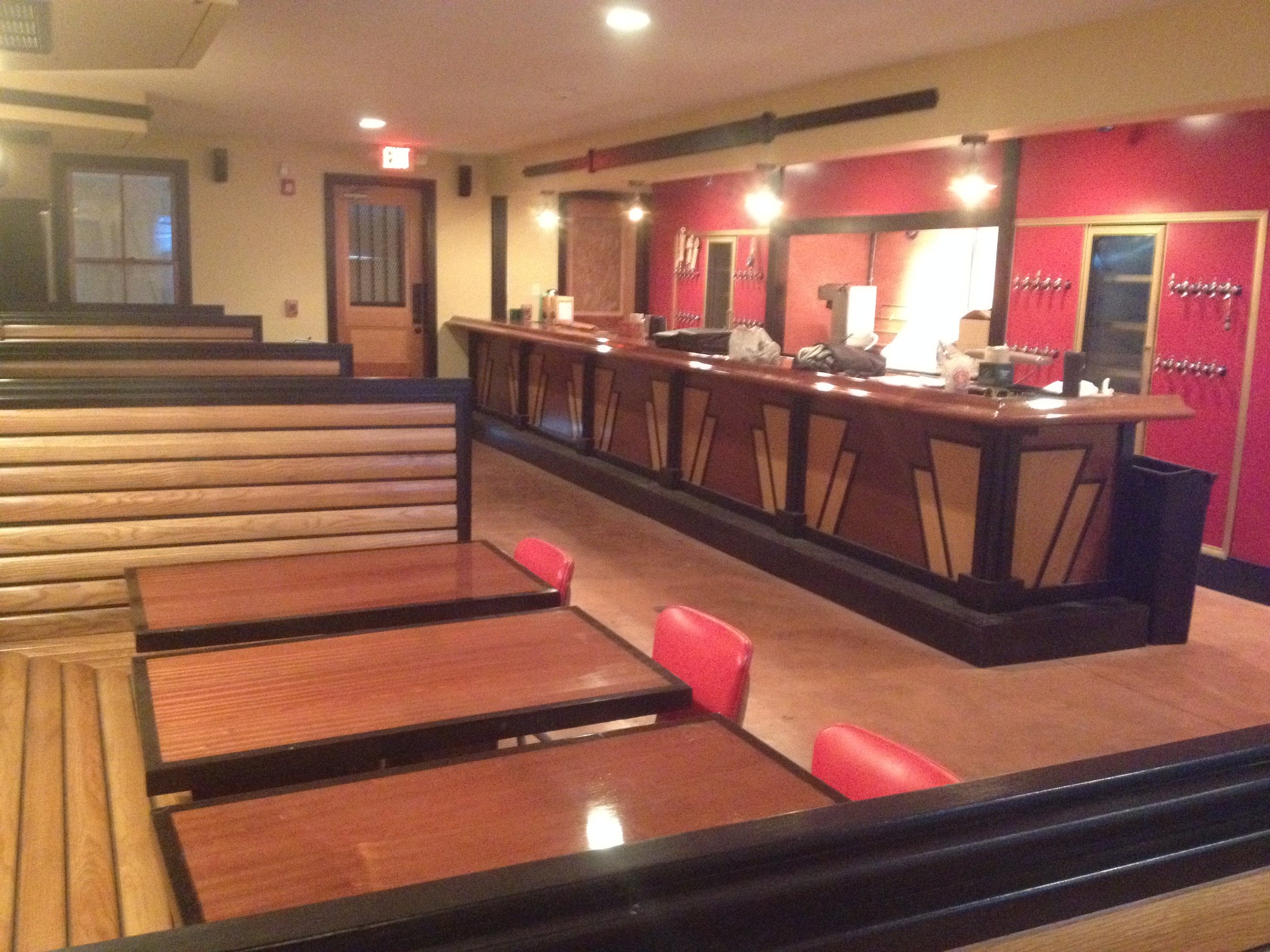 Empty restaurant with wooden tables and red chairs, a bar in the background with taps and kitchen area behind, warm lighting and wood accents.