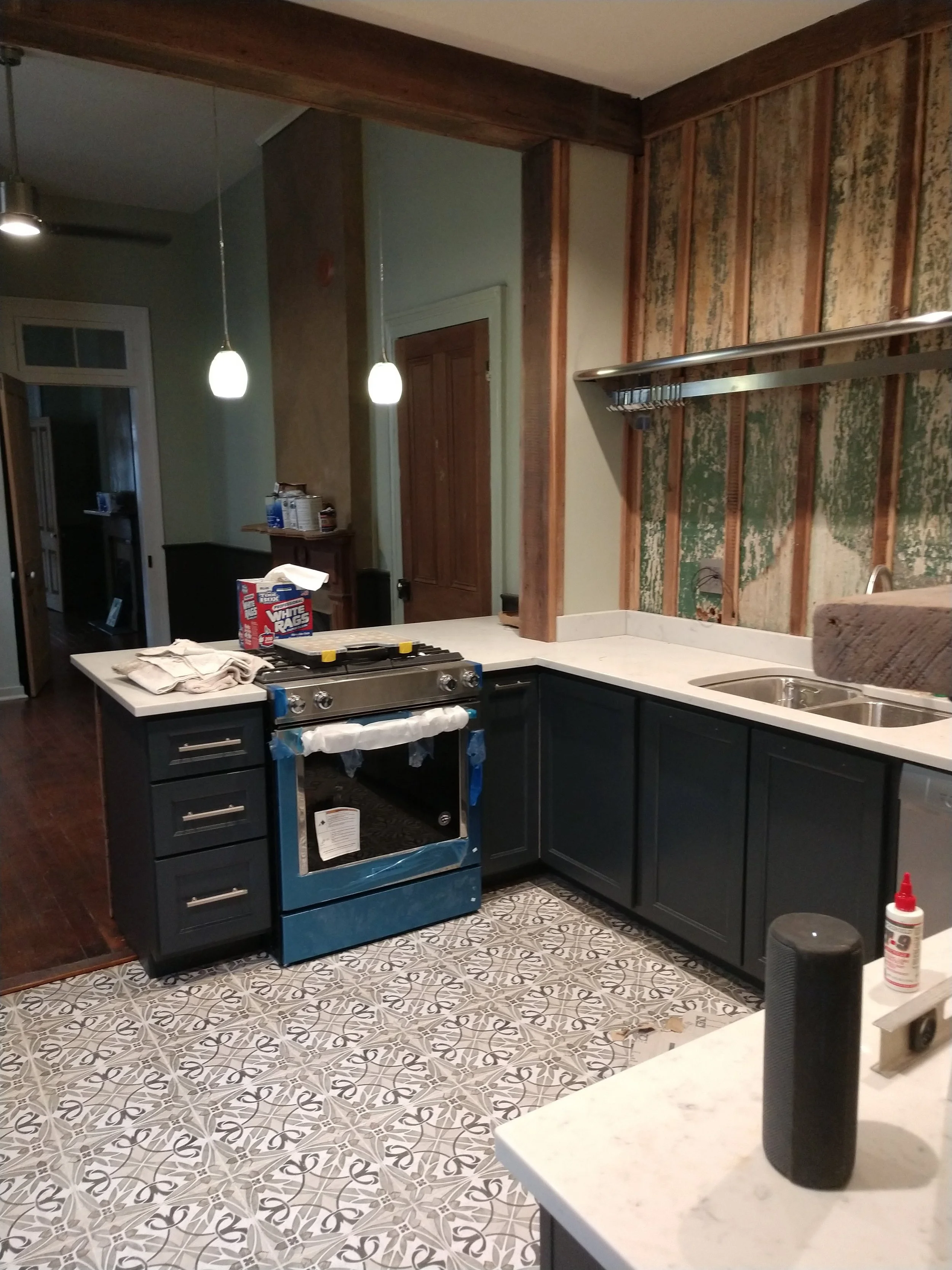 Kitchen with dark lower cabinets, white countertop, double sink, patterned tile floor, stove with plastic cover, partially removed wall with exposed wood framing, hanging shelf, and various items on counters.