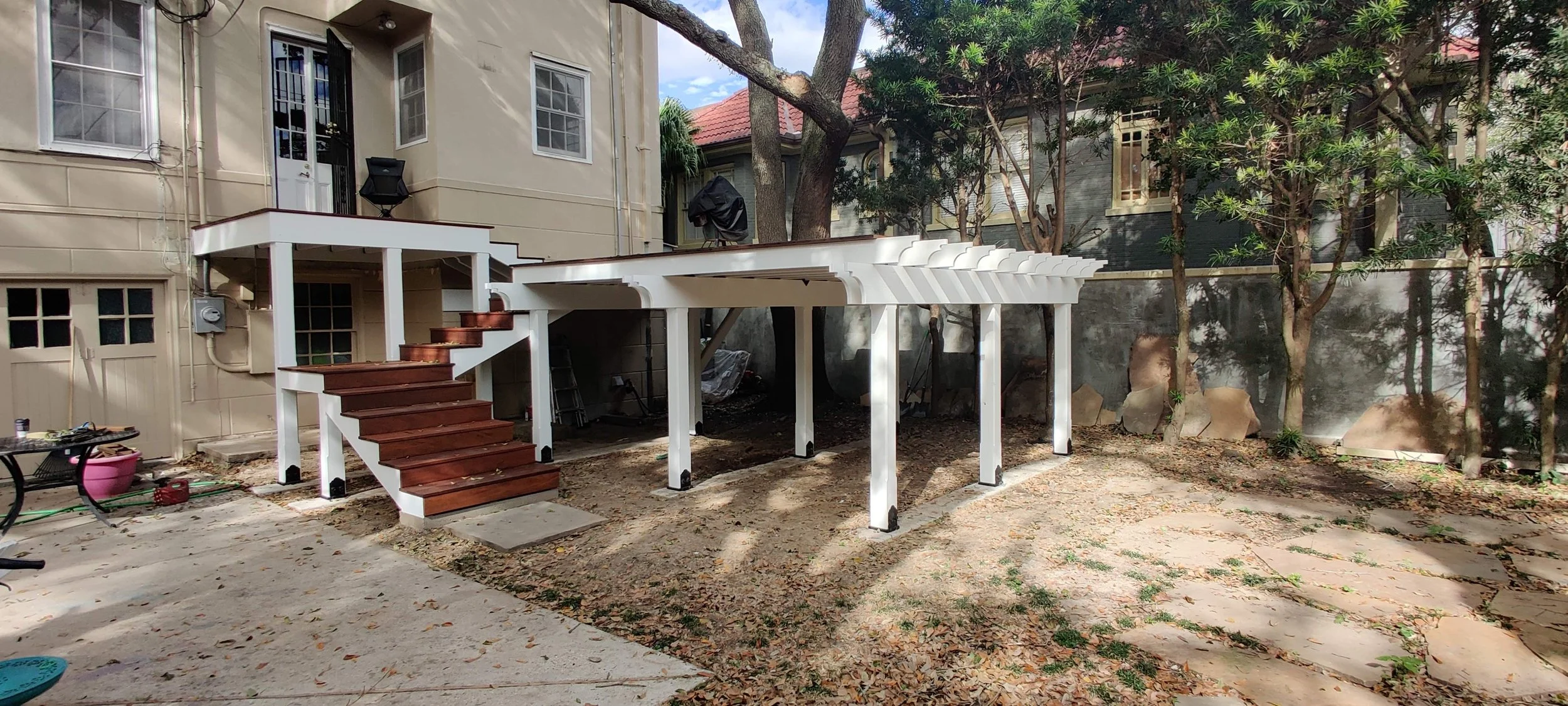 A backyard with a partially built white wooden deck with stairs, next to a beige house with multiple windows. There are trees and a gray wall in the background, and the ground is covered with leaves and dirt.