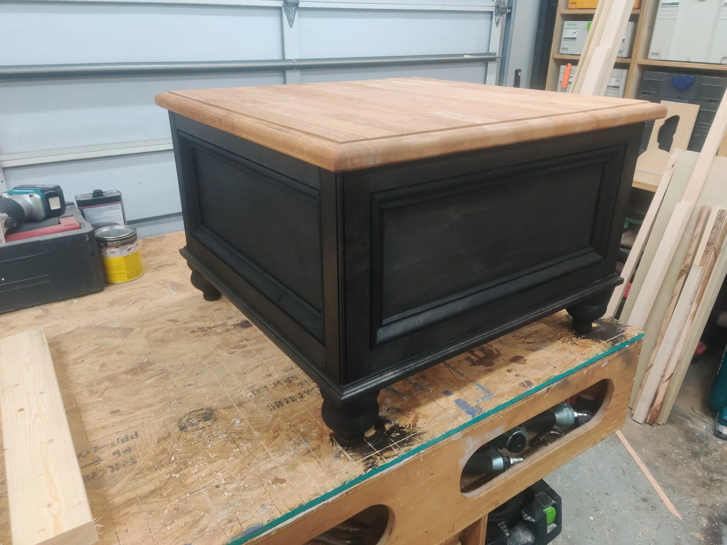 A black and wood-colored wooden storage chest or bench with a natural wood top, placed on a workbench in a woodworking shop.