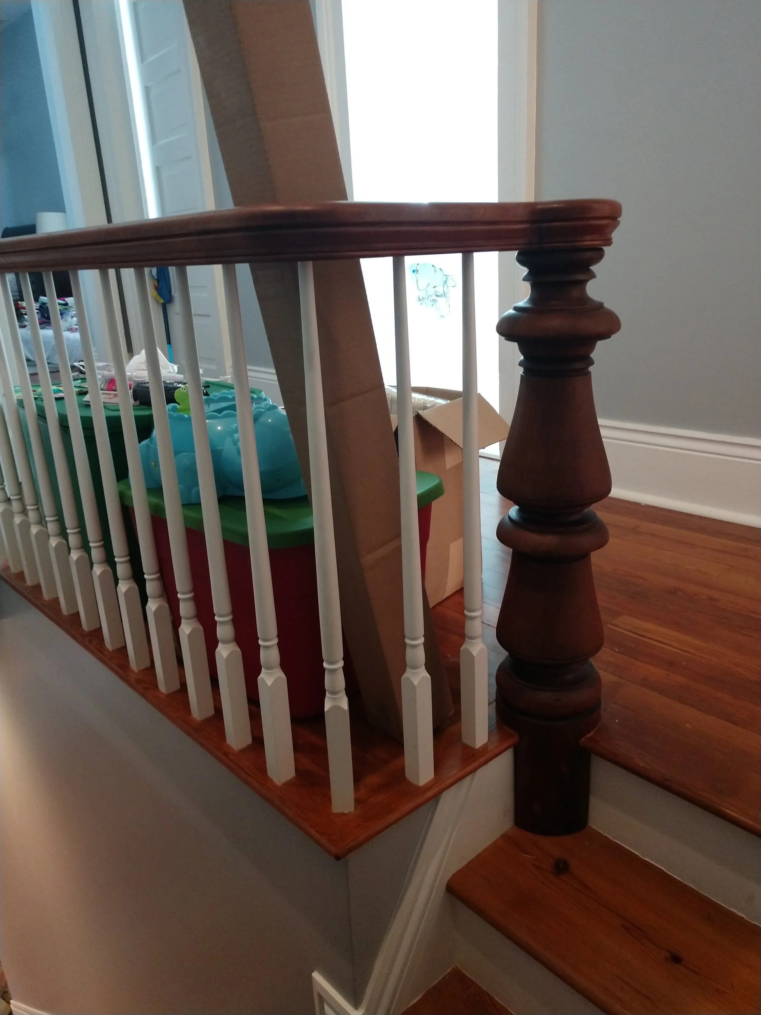 Part of a staircase with a wooden handrail and white spindles. Several cardboard boxes and plastic storage containers are on the upper landing, with some toys visible inside the containers. The background shows a partly open door and a bright window.