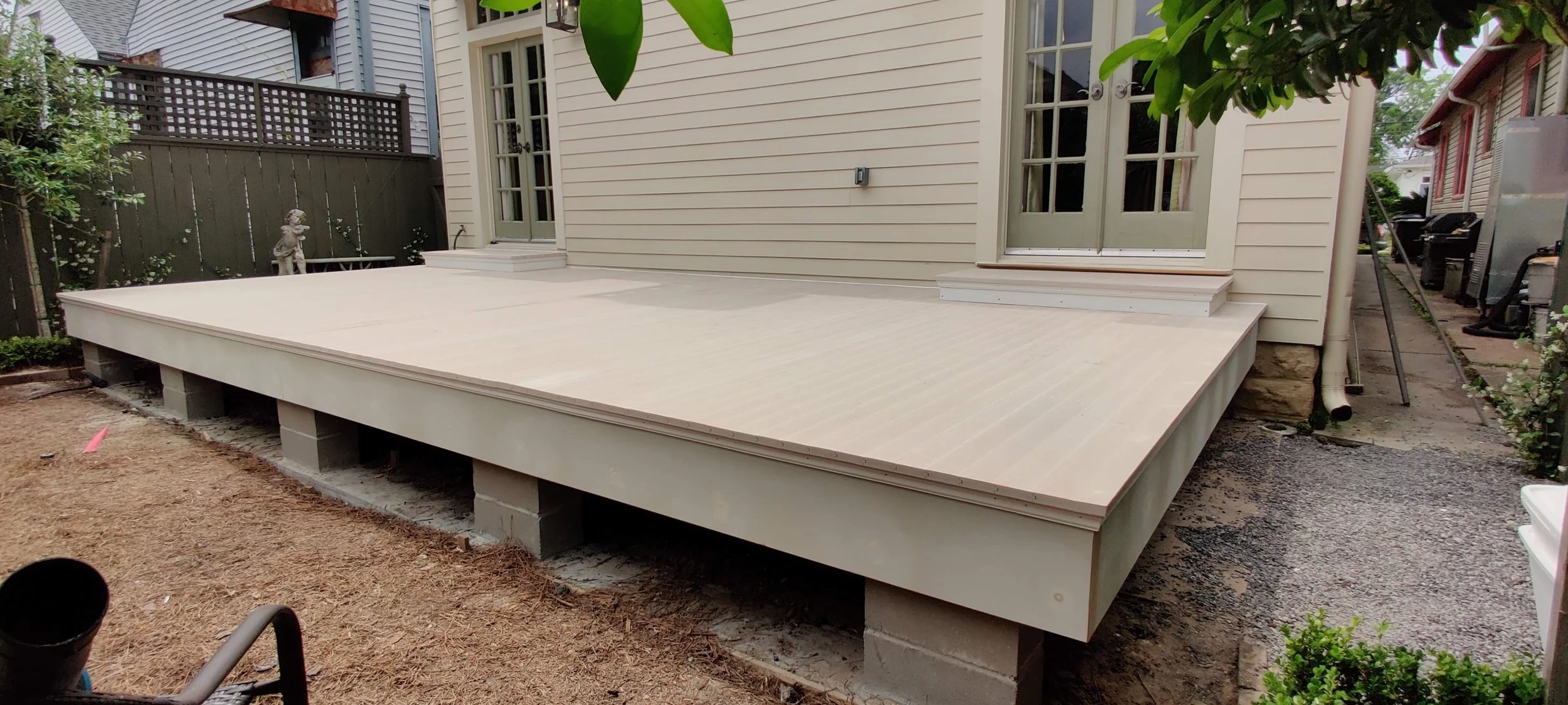 A newly built deck attached to the back of a house, with a sliding glass door leading to the interior. The deck is elevated and supported by concrete blocks, and there is a gravel pathway along the side of the house.