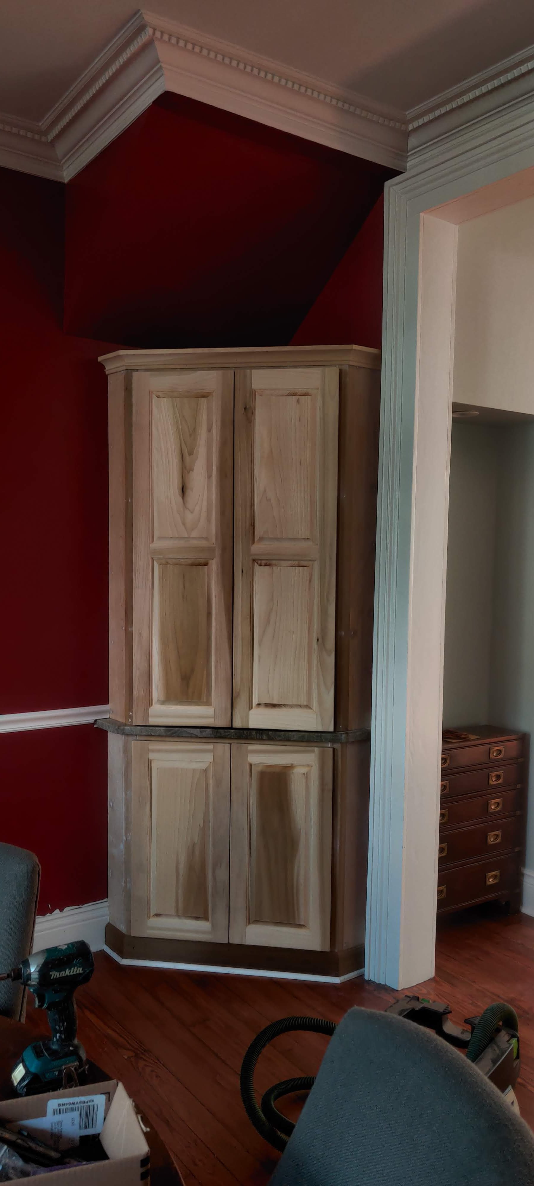 A corner cabinet with four wooden doors in a room with dark red and beige walls and detailed crown molding.