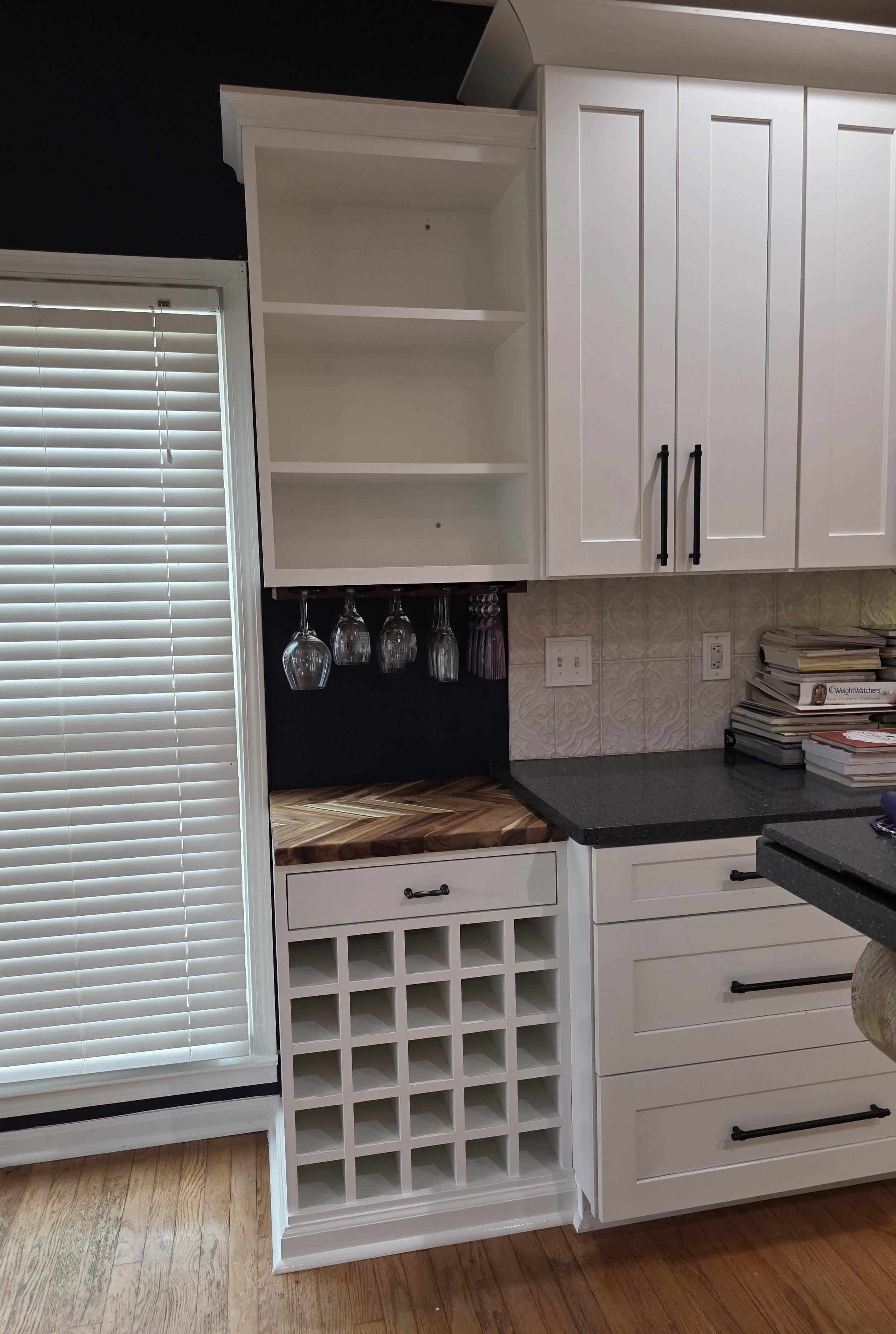 Kitchen cabinet with open shelves, hanging wine glasses, wine rack, countertop, and stacks of books.