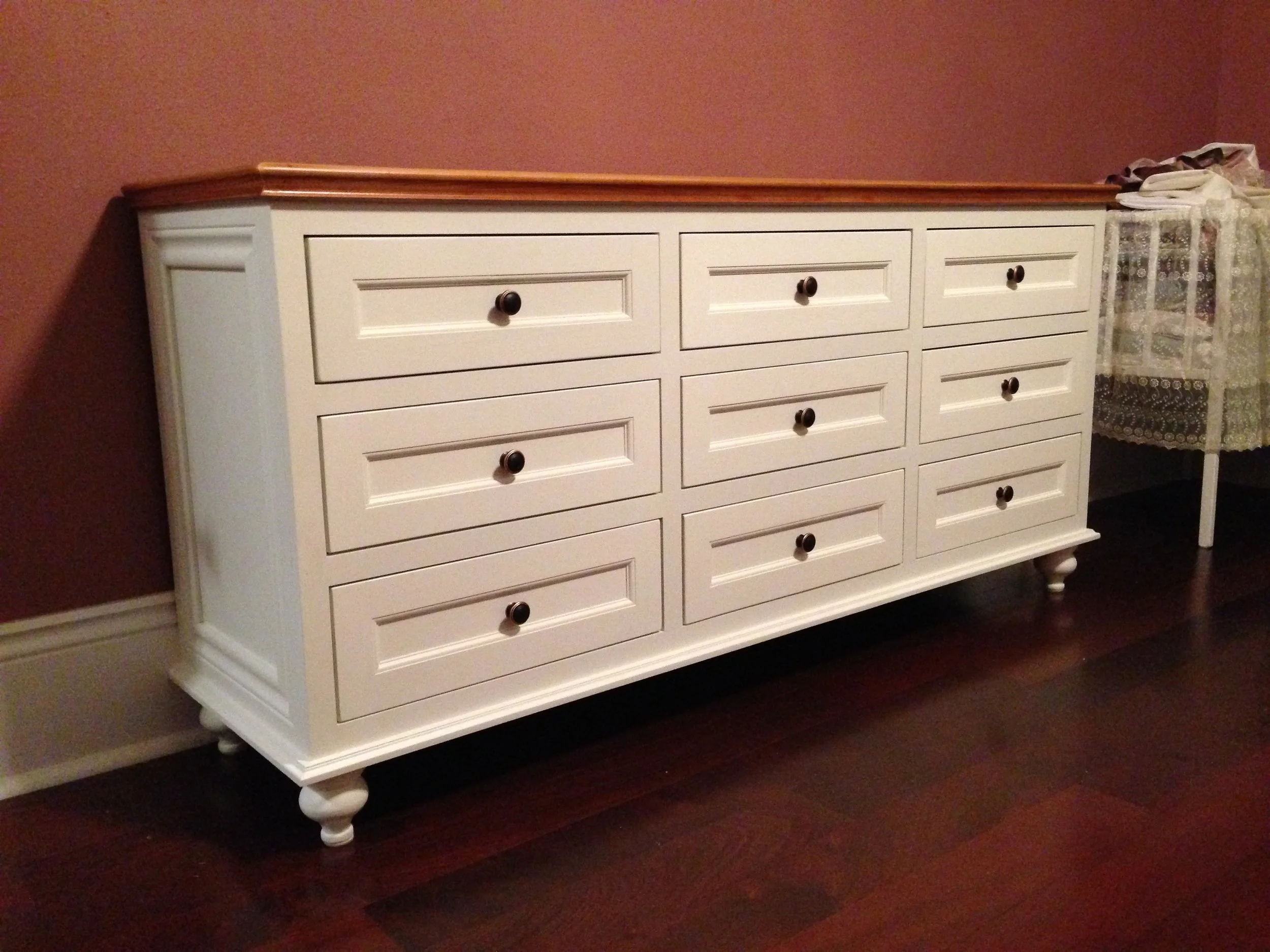 White wooden dresser with nine drawers, black knobs, and a wooden top, located against a pink wall, with a lace-covered table to the right.