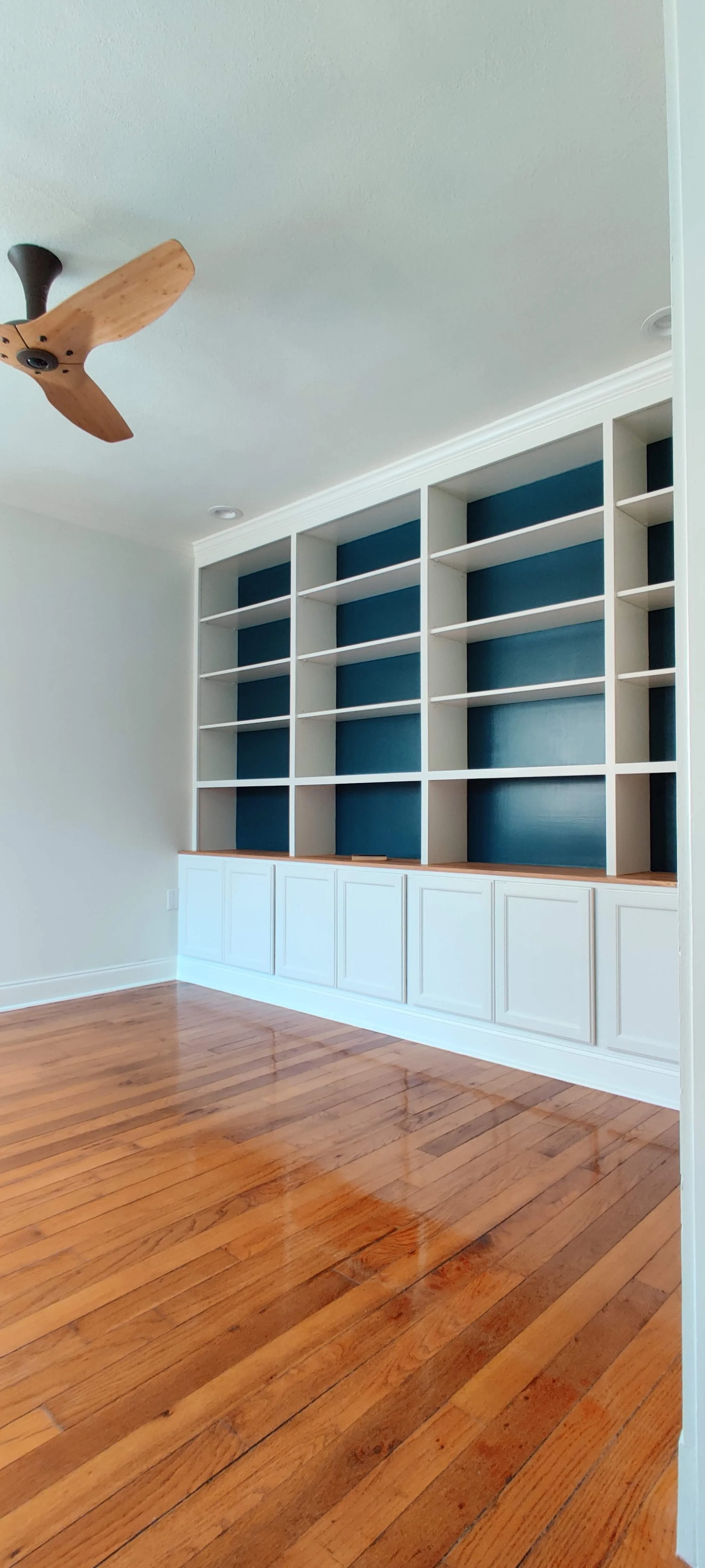 Empty built-in bookshelf with a blue back panel in a room with hardwood floors and a ceiling fan.