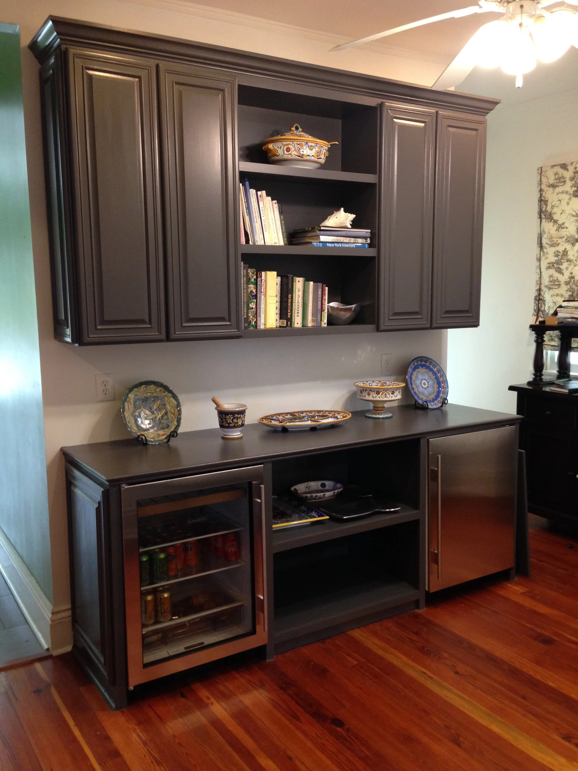 Built-in dry bar with an open shelf in the center filled with books and decorative objects, a countertop below holding decorative plates and bowls, a small fridge with drink cans, and a hardwood floor.