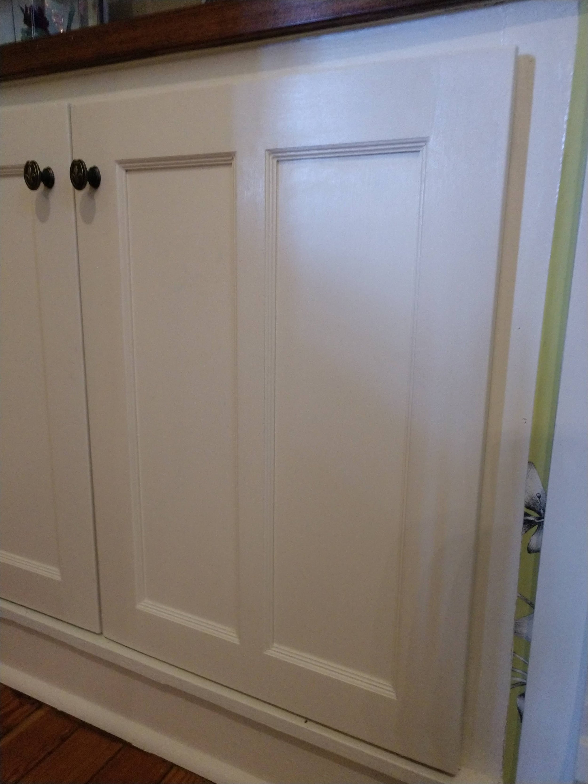Close-up of cream-colored kitchen cabinet with dark round knobs and decorative trim, partially revealing a butterfly-themed wallpaper behind.
