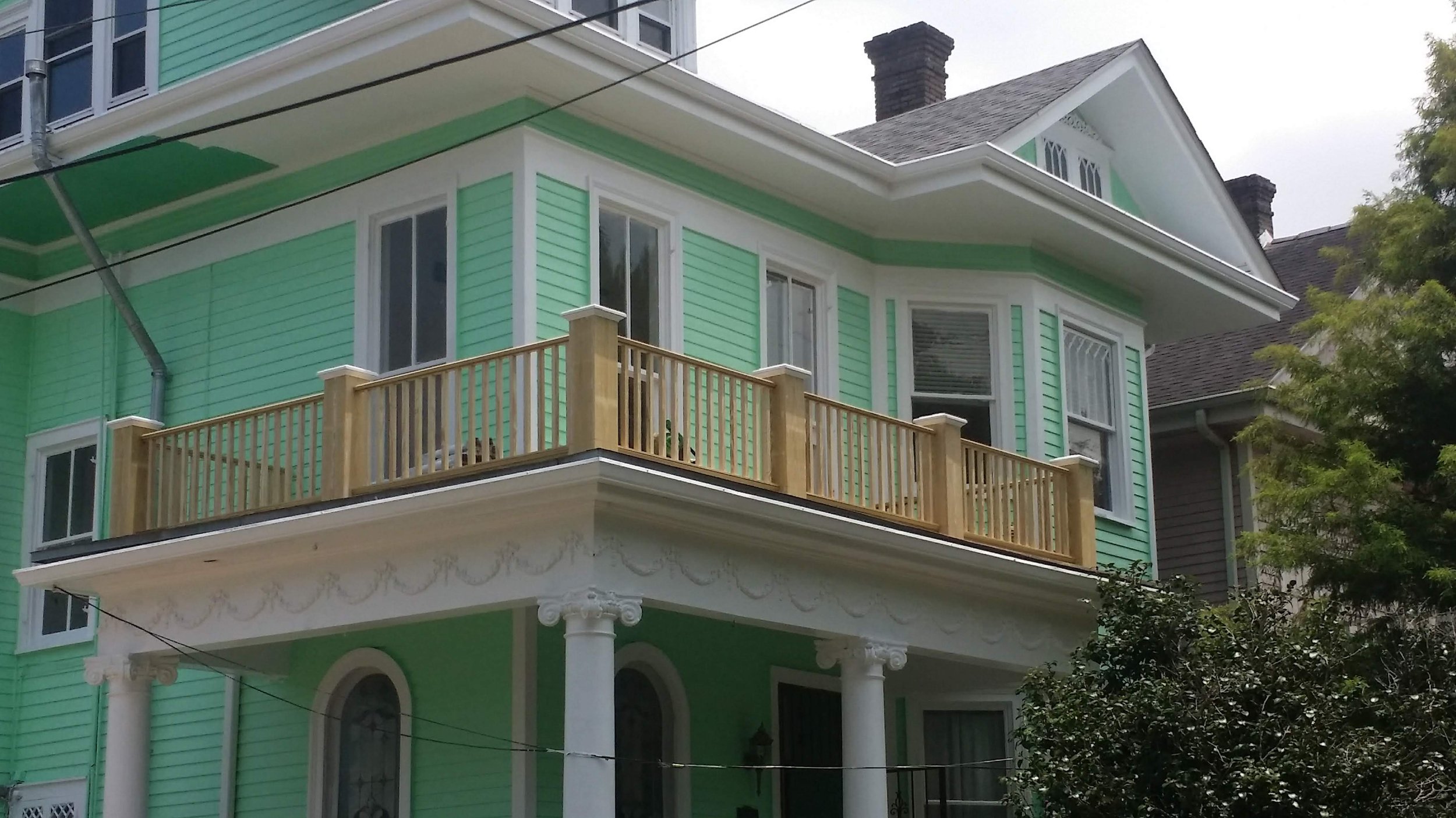 A multi-story house painted in light green with white trim, featuring a wooden balcony railing on the second floor, decorative columns supporting the porch, and a sloped roof with multiple windows.