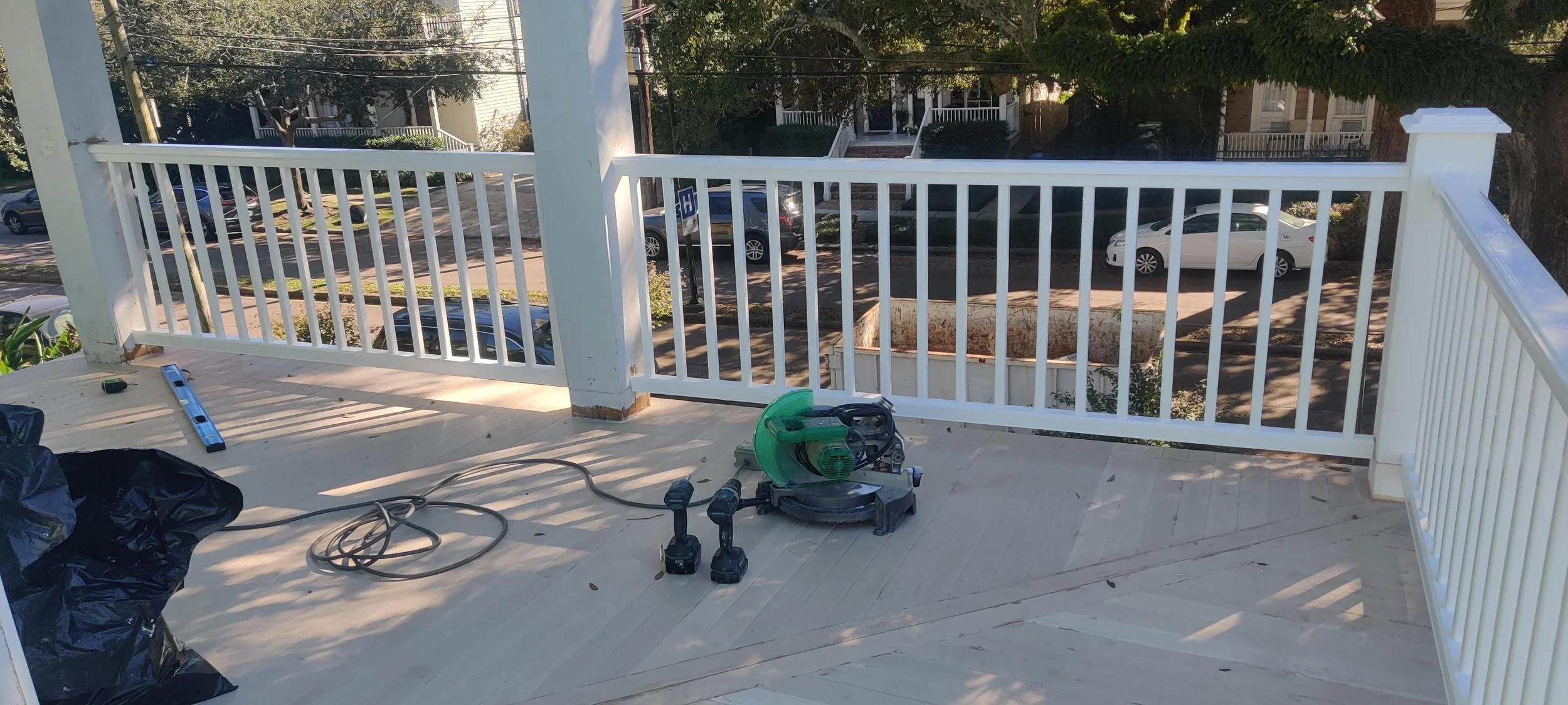 On a porch, there are construction tools including a circular saw with a green blade guard, two cordless drills, a level, an extension cord, and a black plastic bag, with a painted white railing in the background overlooking a residential street with