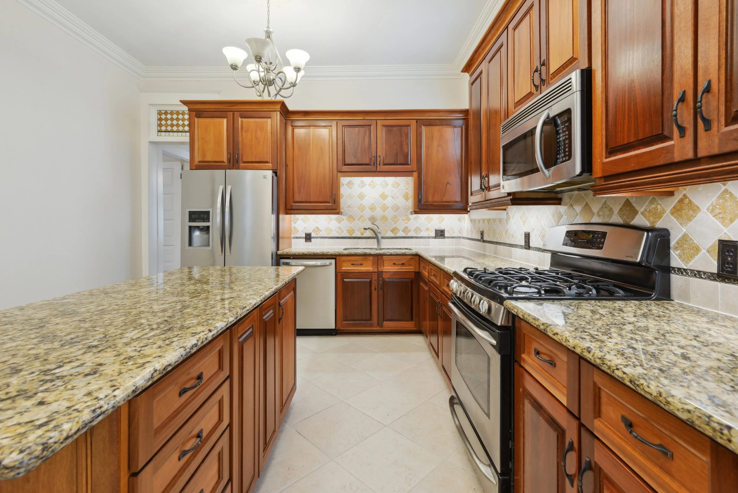 Kitchen with mahogany cabinets, granite countertops, stainless steel appliances including a microwave, gas stove, refrigerator, and a small dishwasher, beige/tan tiled walls, tiled floor, and a ceiling light fixture.
