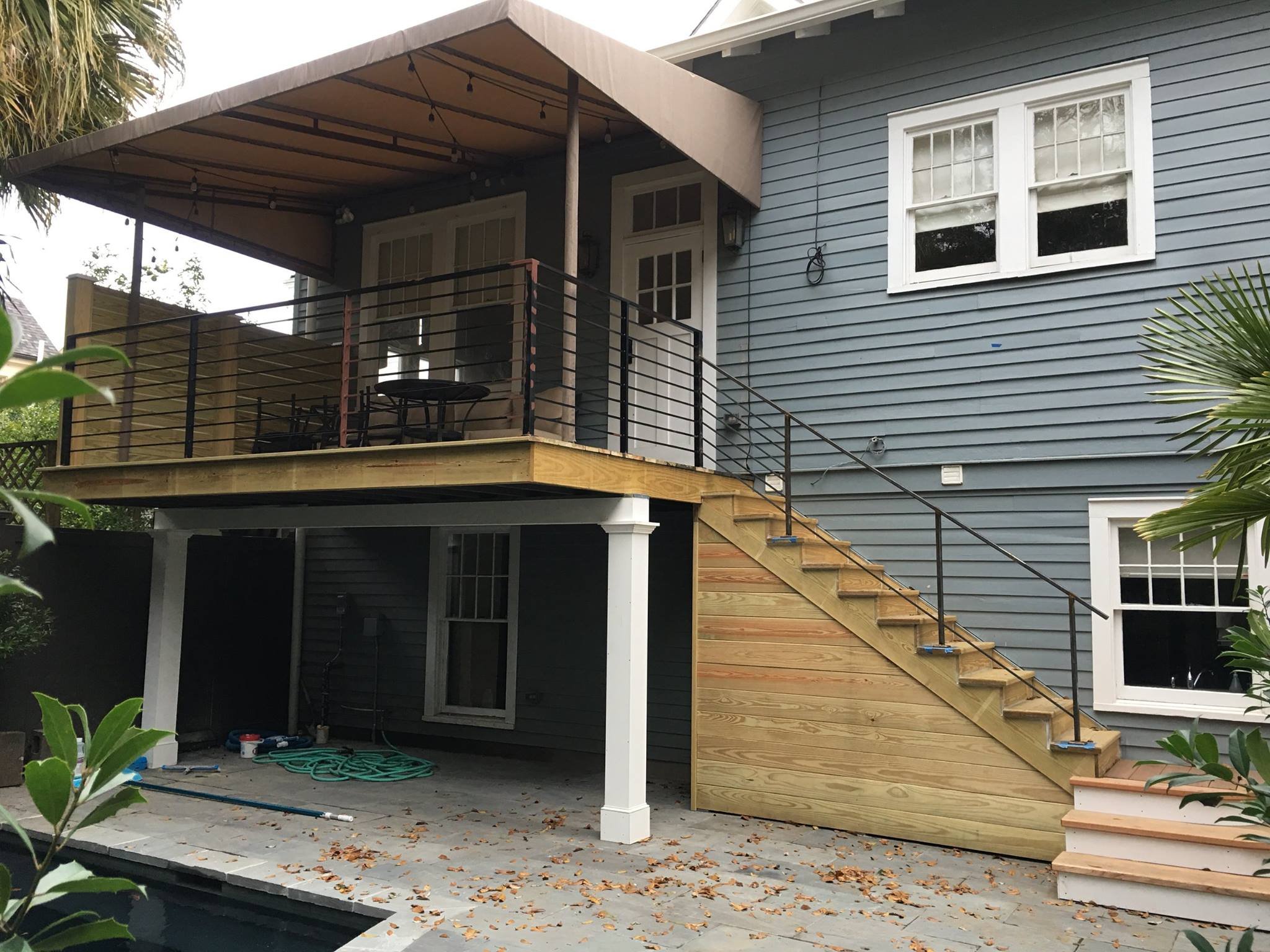 A two-story house with an outdoor balcony on the upper level, featuring a wooden deck, black railing, and string lights. The house has blue siding, white trim around windows, and stairs leading up to the balcony.