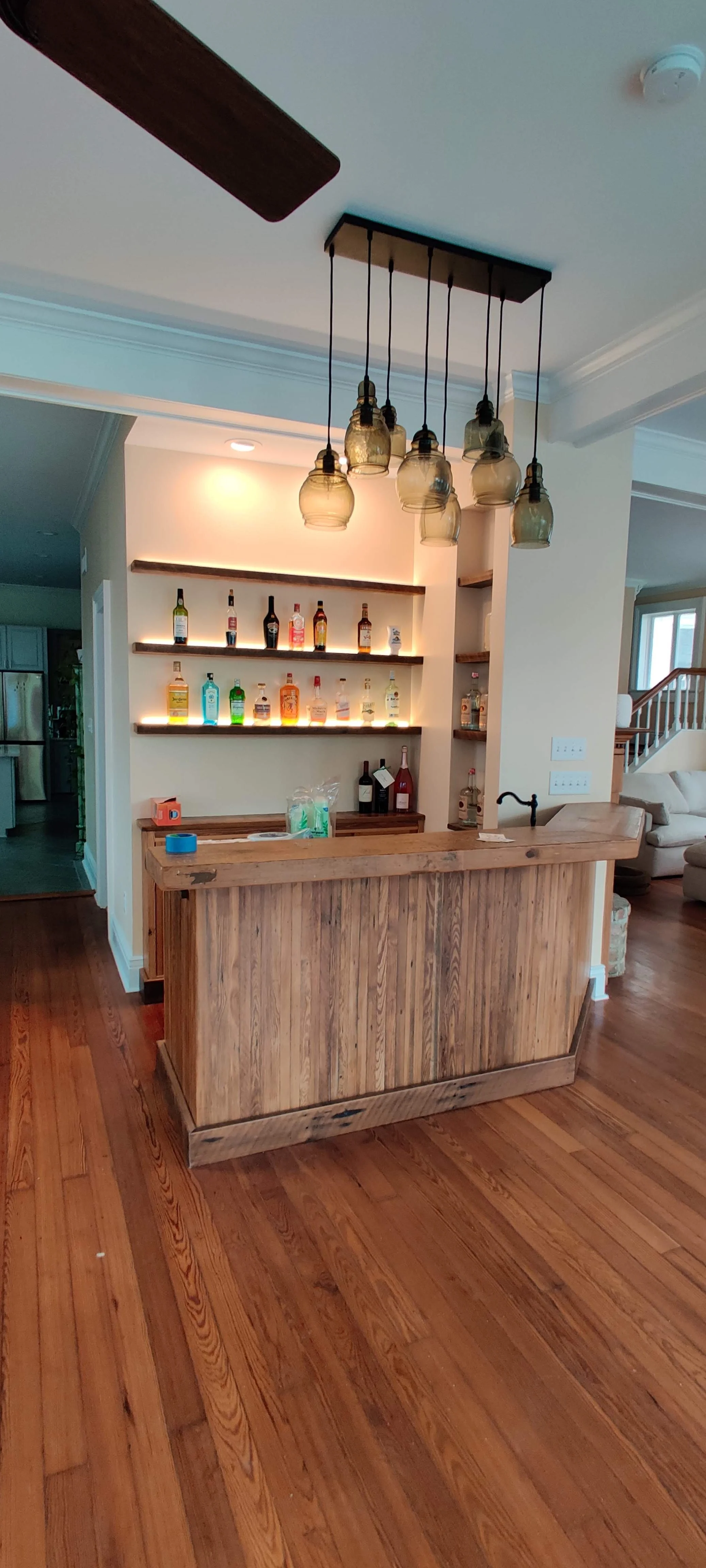 A home bar with a wooden counter and shelves displaying bottles of alcohol, a hanging glass pendant light fixture, and hardwood flooring.