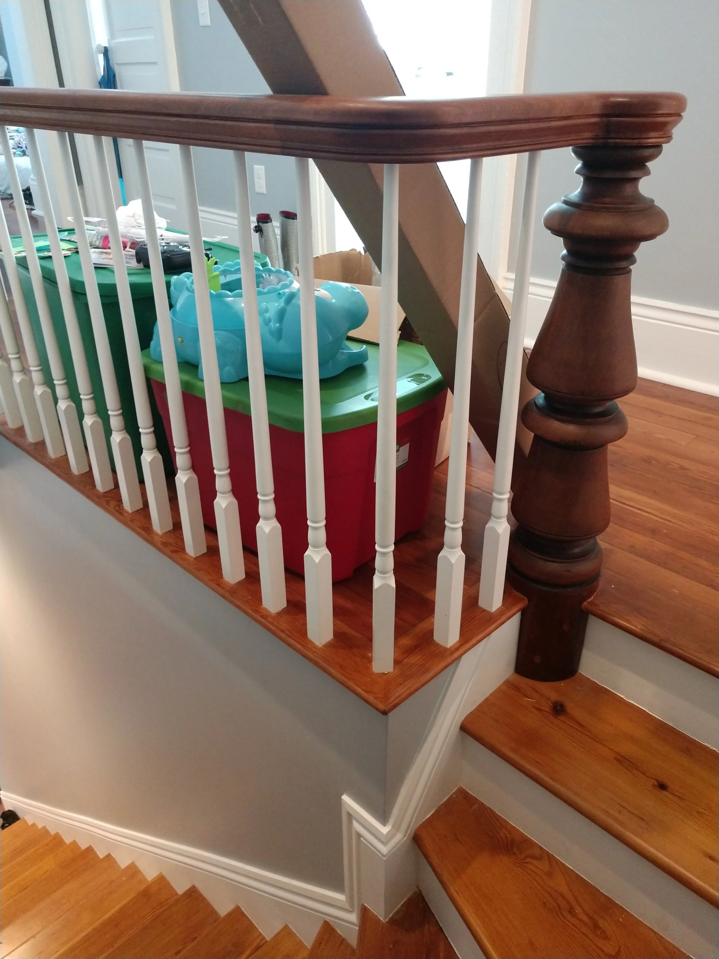 View of a staircase landing with a large red and green plastic storage bin containing a toy crocodile, placed behind white railing spindles and a wooden newel post.